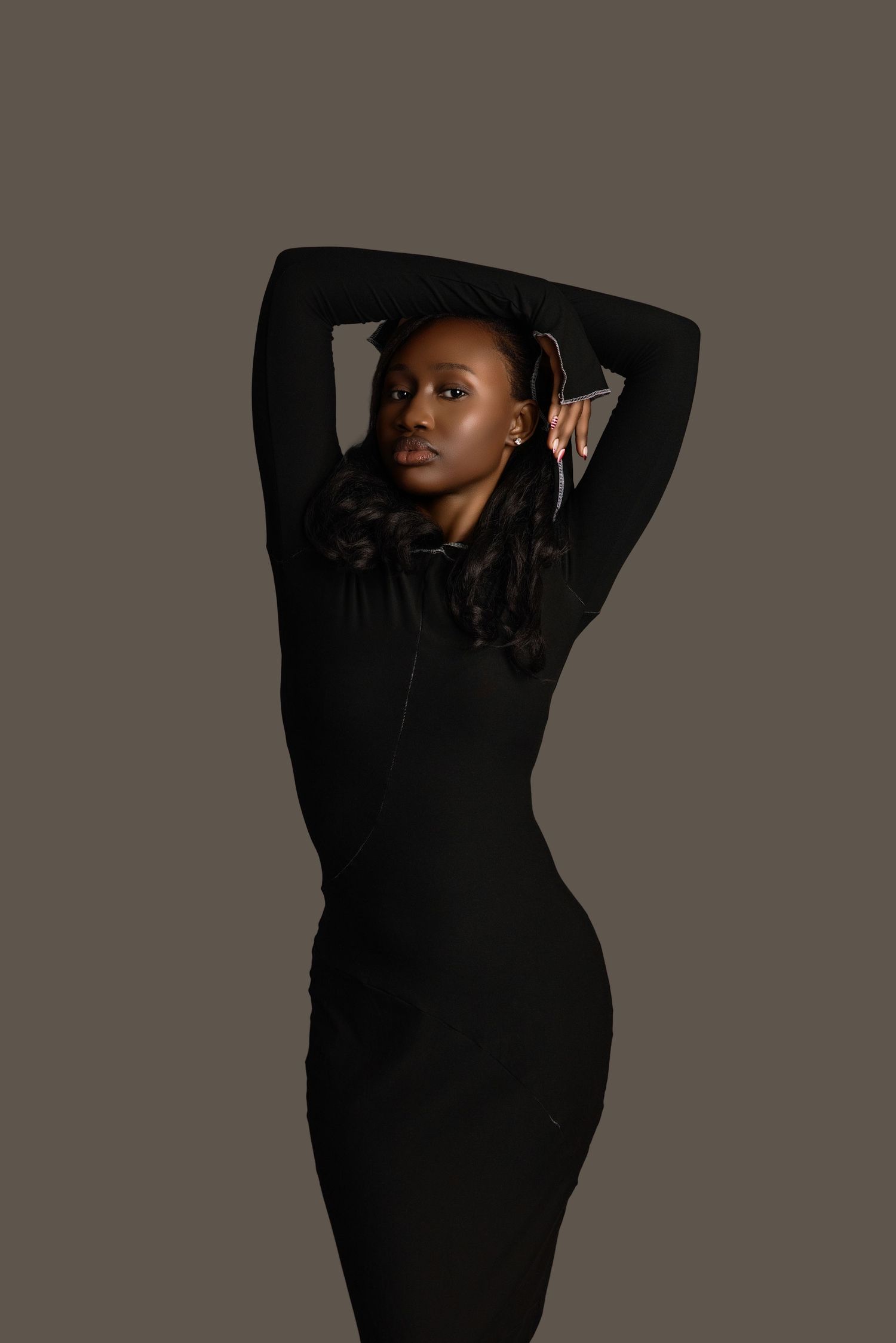 Black female model in black zip-front midi dress with arms raised overhead, studio portrait.