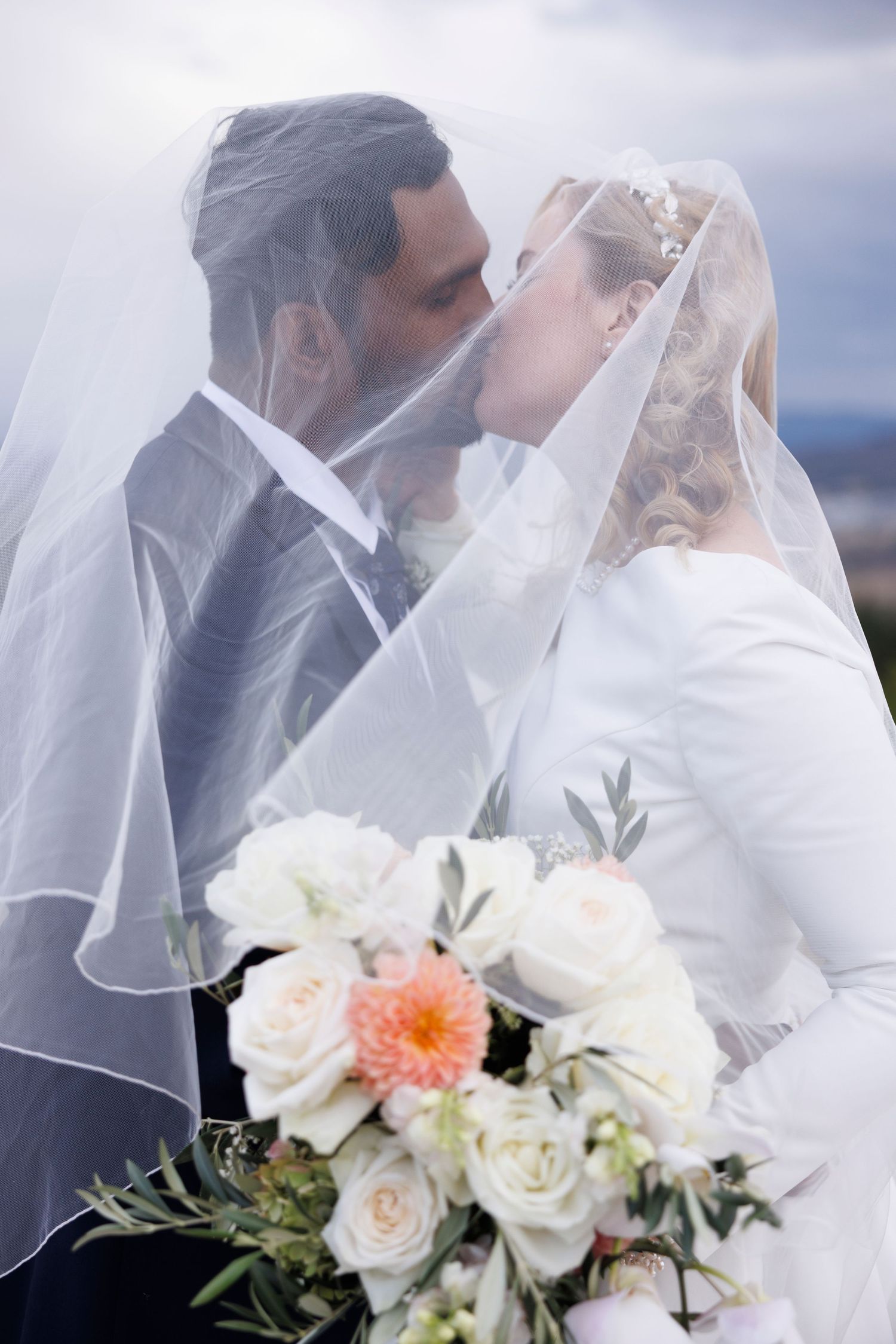 Bride and groom kissing under a sheer veil with bridal bouquet in foreground.