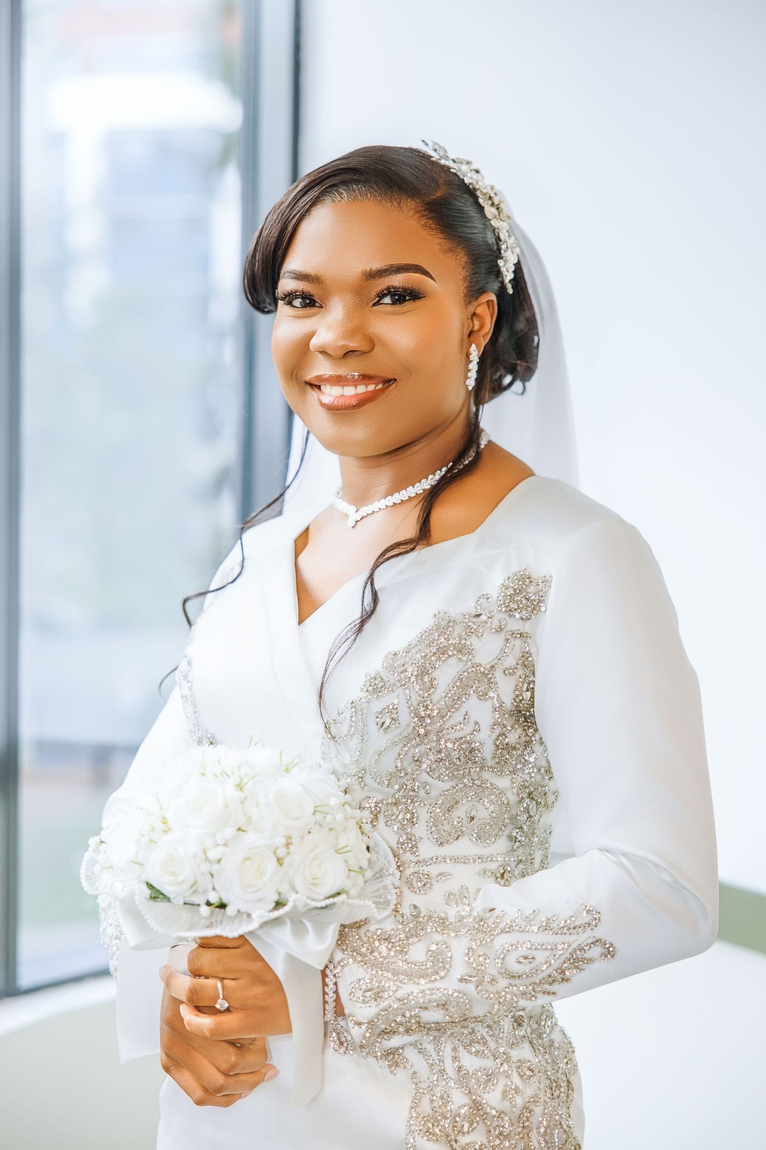 Smiling Black bride in white beaded gown holds bouquet, wearing pearl necklace and crystal tiara with veil.