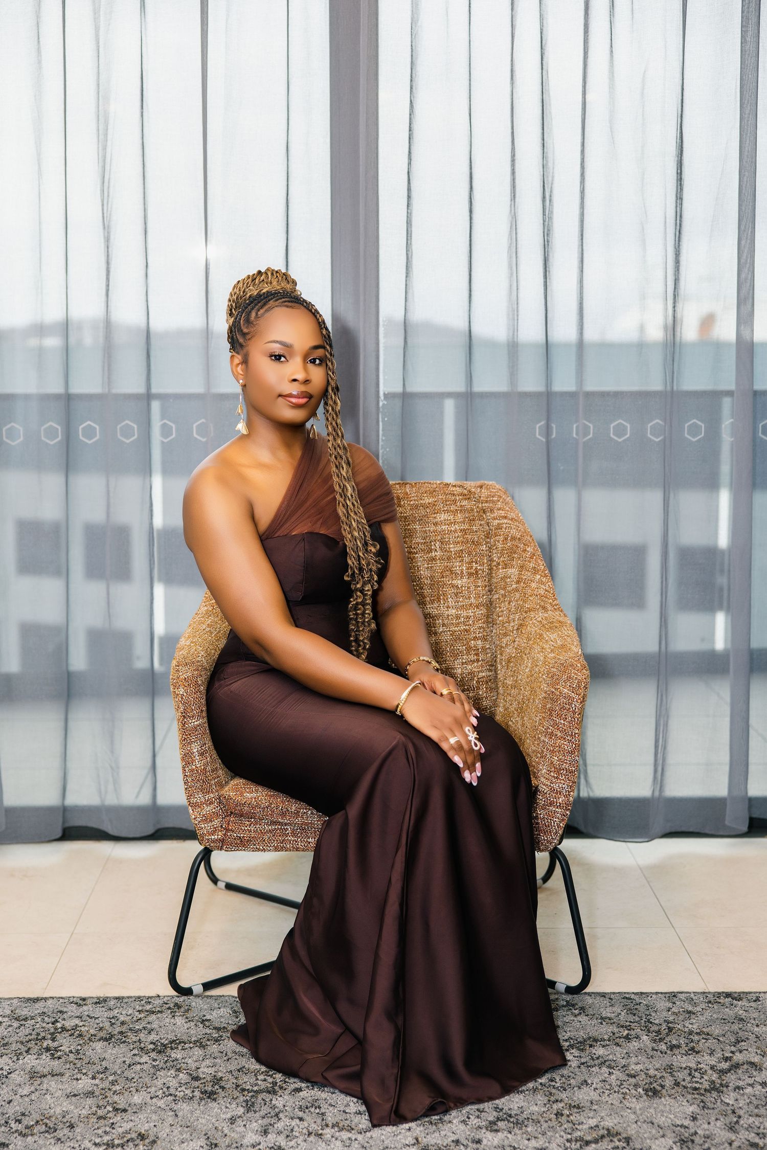 A woman with braided hair in a flowing brown one-shoulder formal gown sits elegantly in a wicker chair.