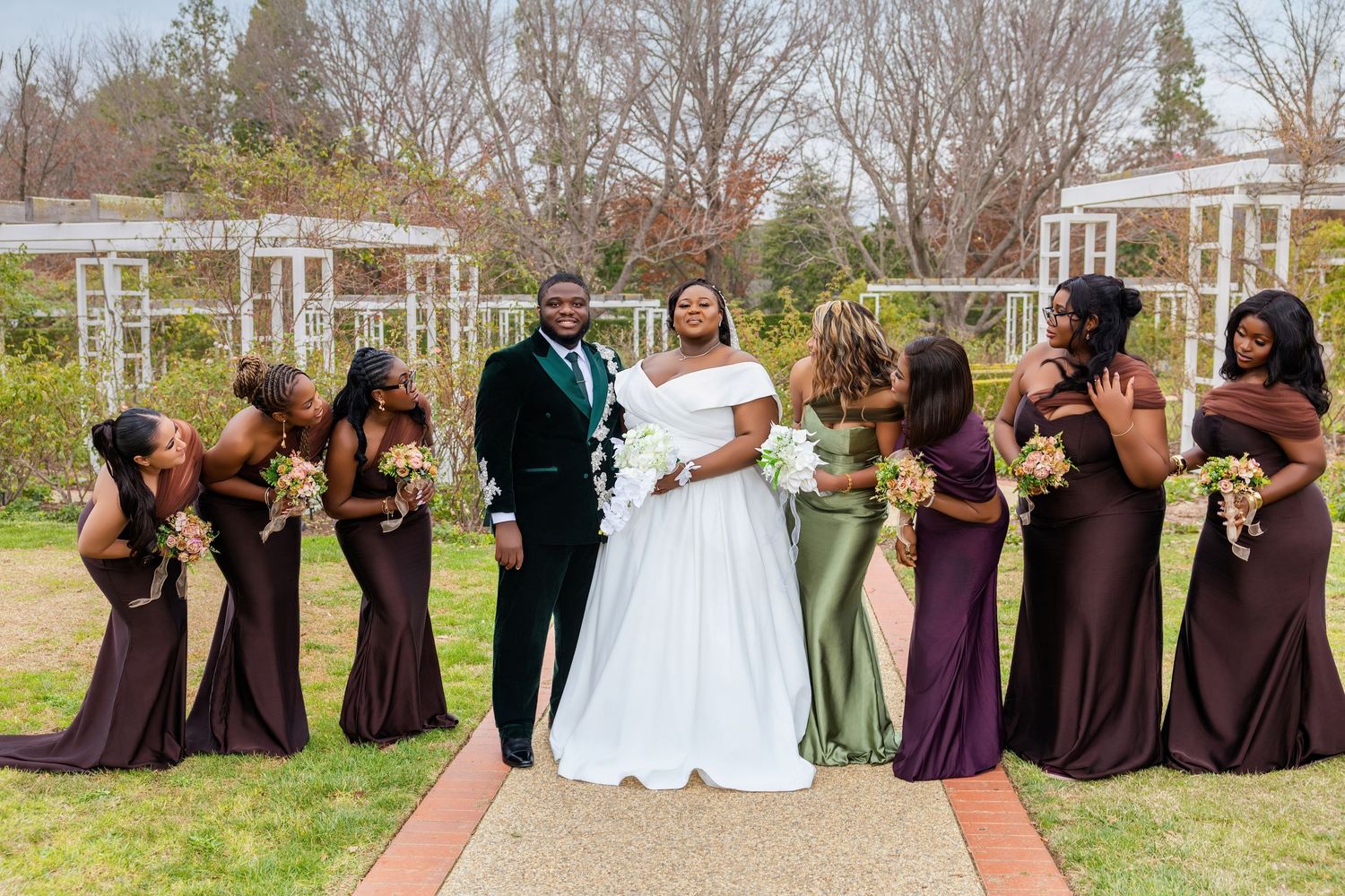 Bride and groom surrounded by bridesmaids in brown and purple gowns at an outdoor garden wedding.