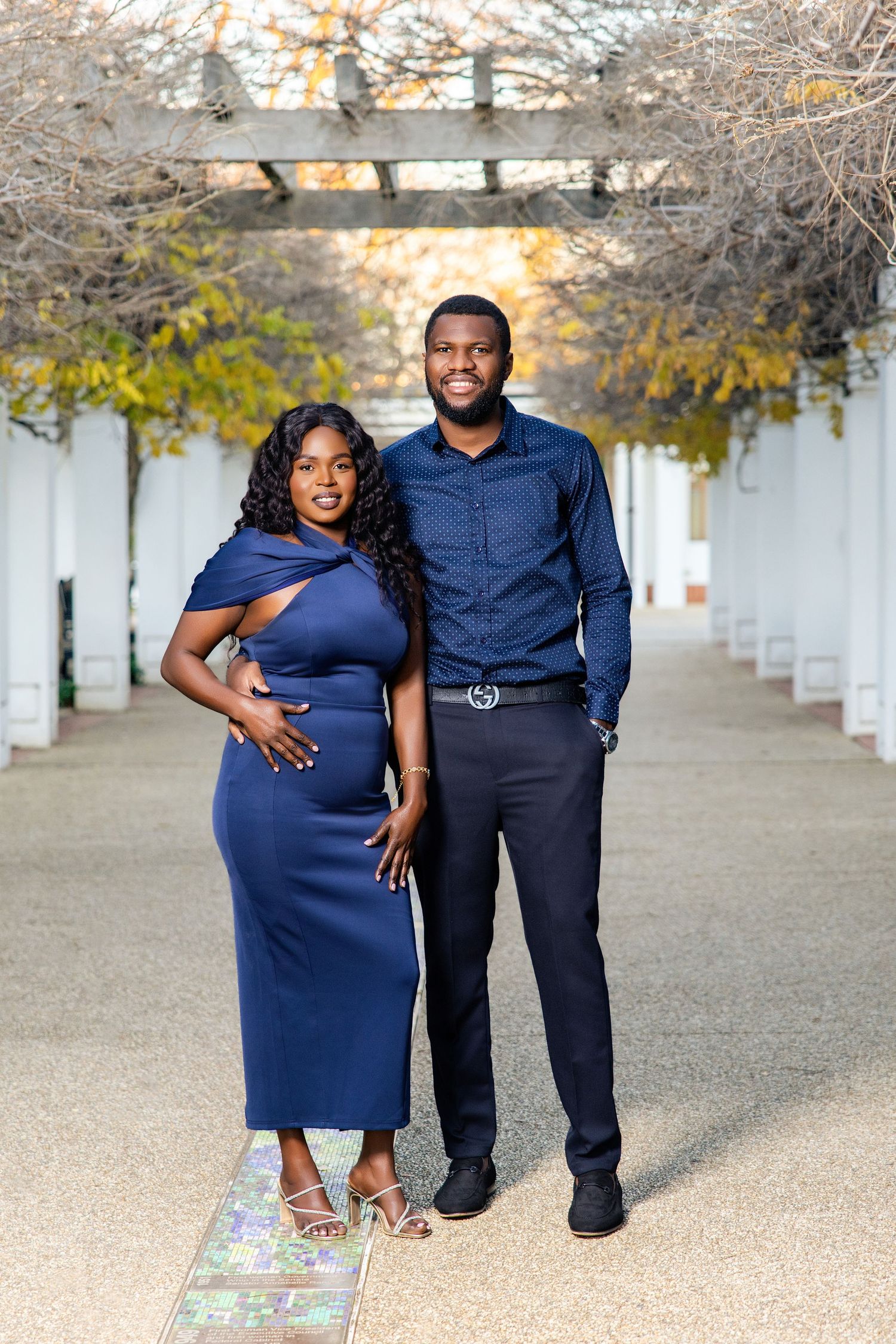 Couple in matching navy blue outfits posing elegantly under a pergola walkway with autumn foliage.