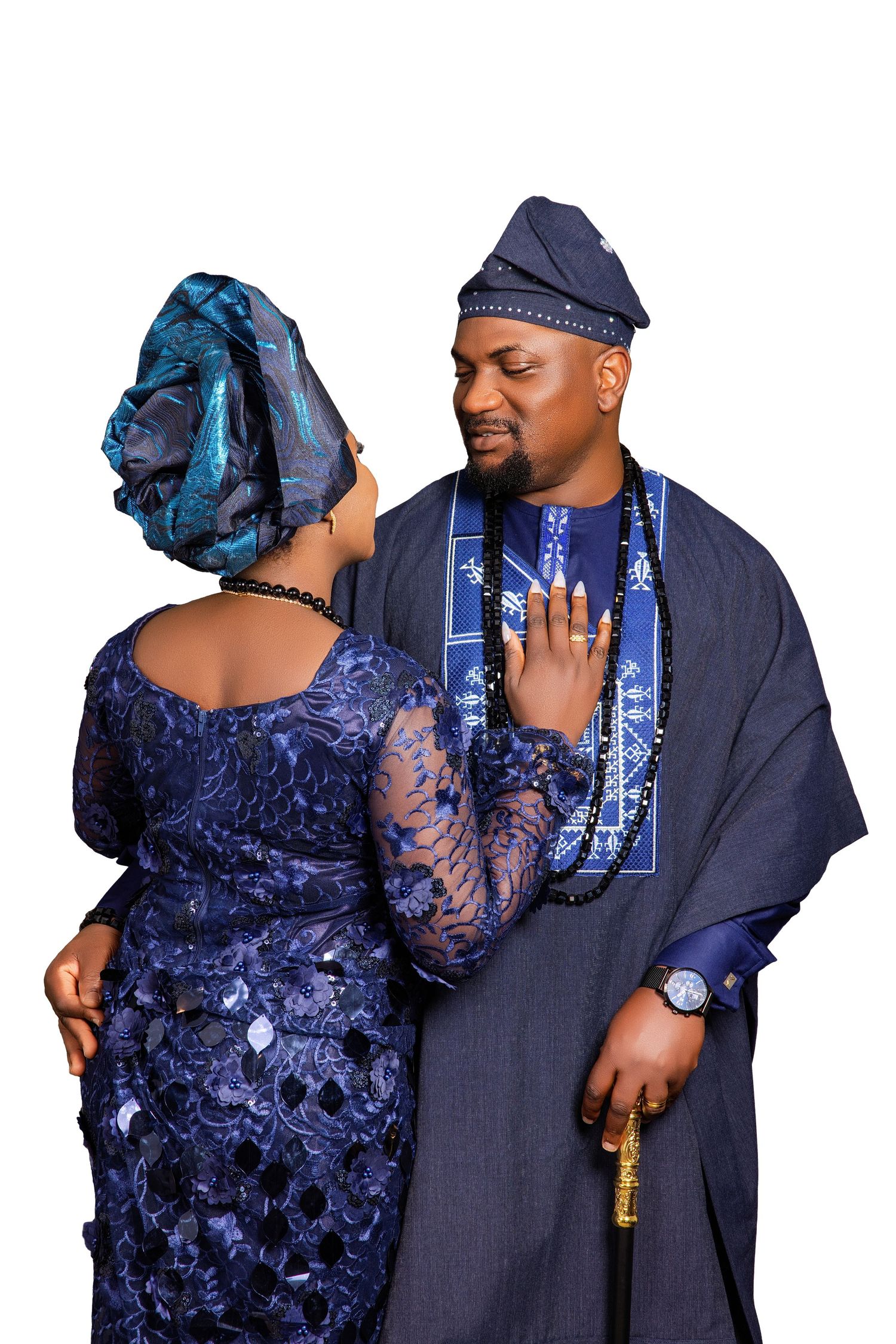 Nigerian couple in matching blue traditional Yoruba attire, agbada and gele, posing together against white background.