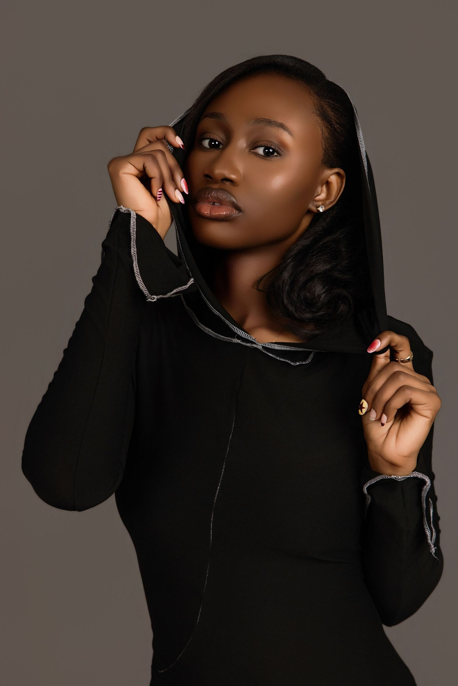 Young Black woman in black hooded dress adjusting hood, close-up studio portrait on gray.
