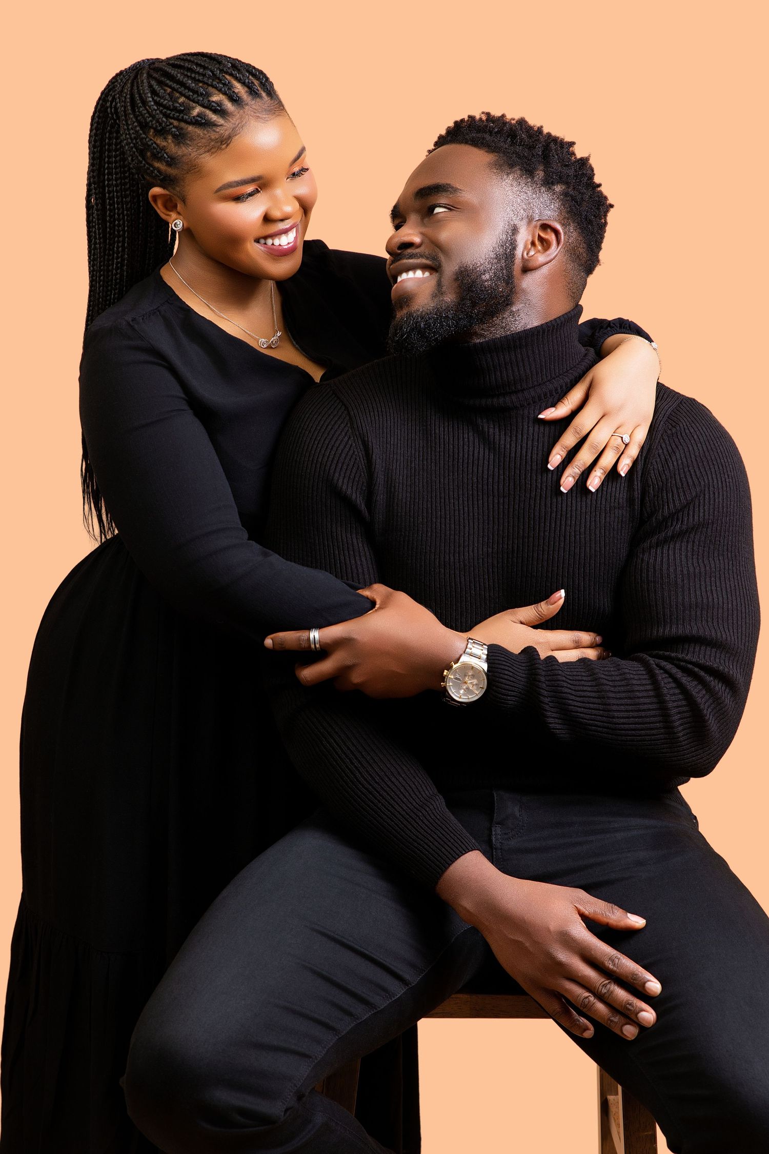 Joyful Black couple in black outfits laughing together, woman standing behind seated man on beige background.