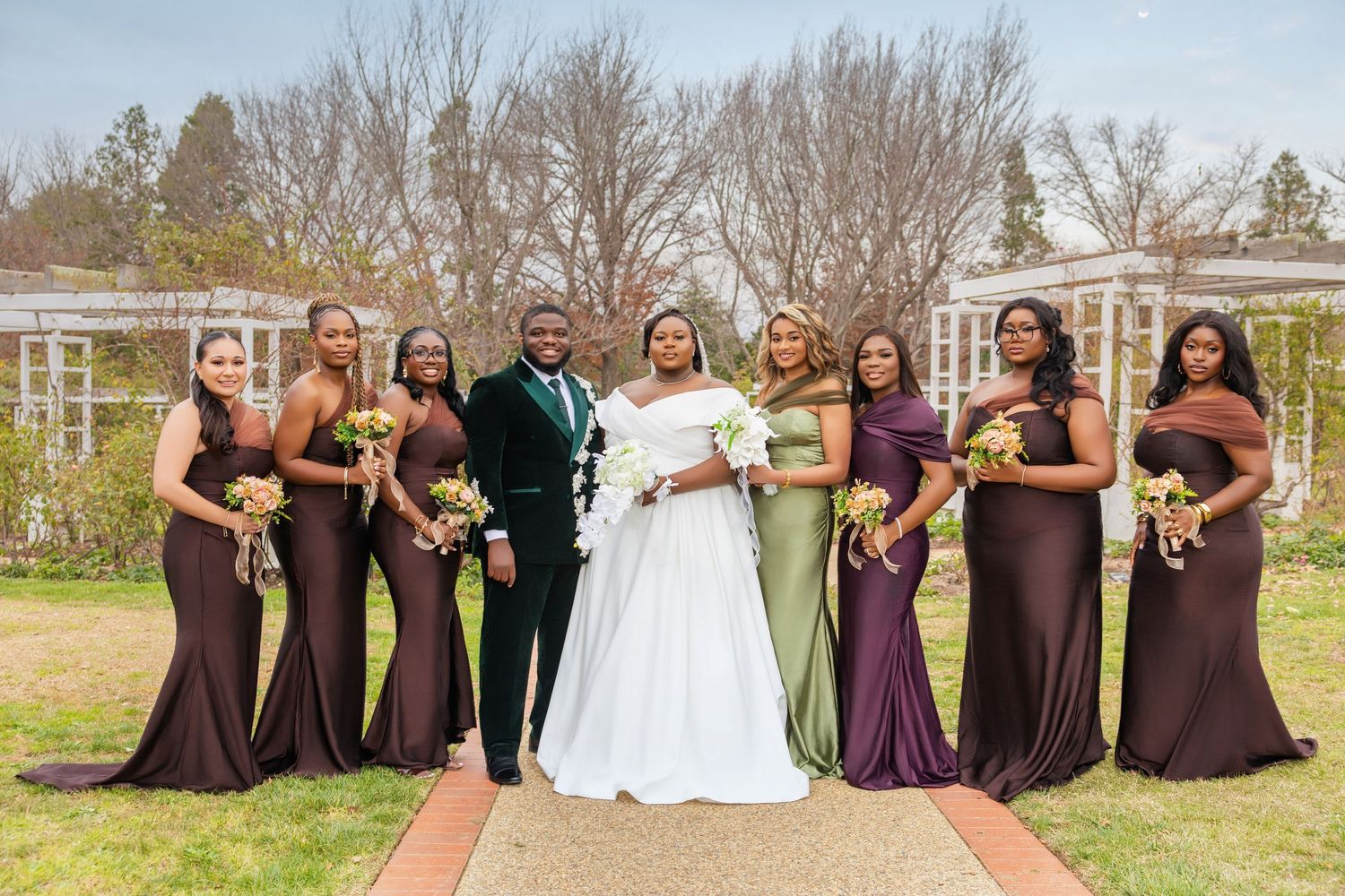 A bride and groom pose with bridesmaids in brown and green gowns in an outdoor garden setting.