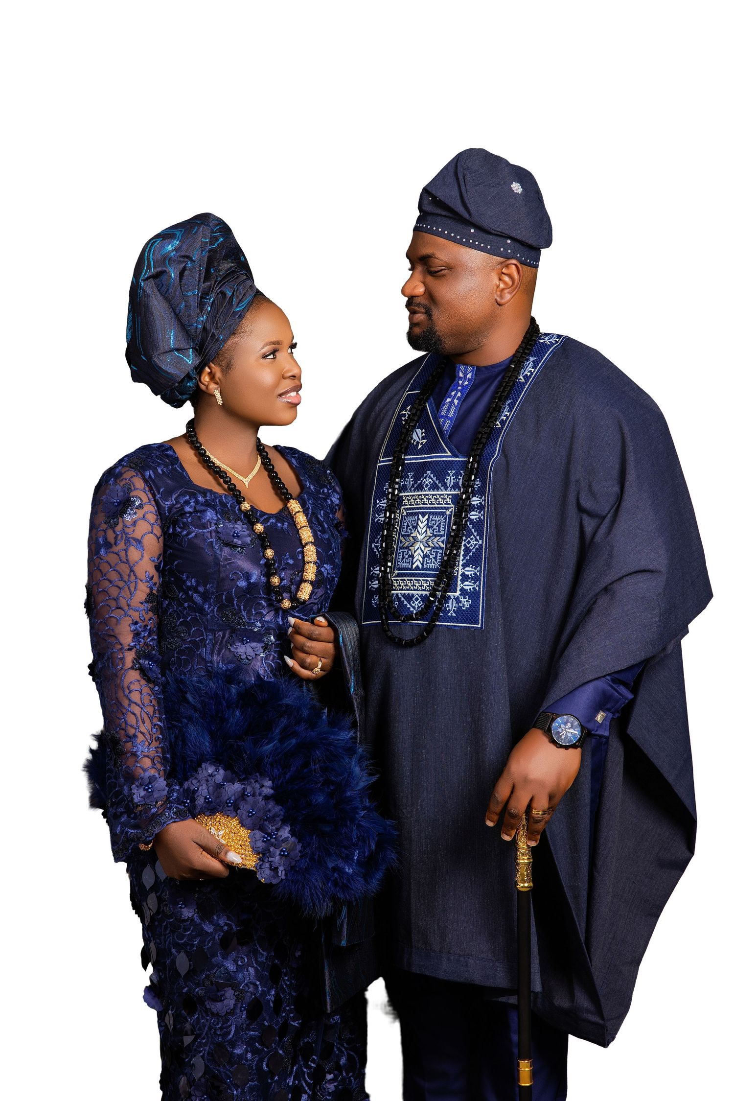 Nigerian couple in traditional Yoruba attire, wearing matching navy blue outfits with gold beaded jewelry, posing together.