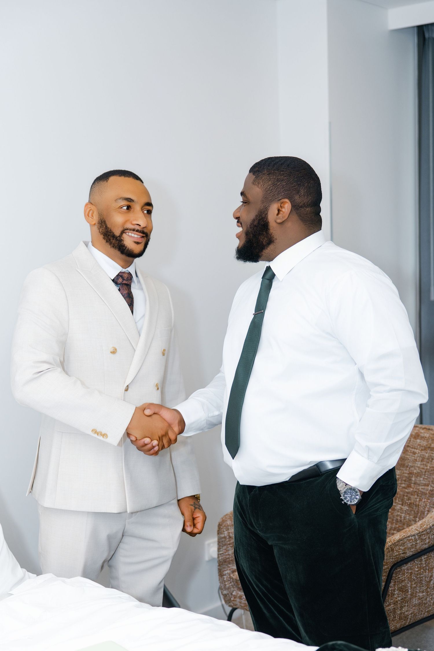 Two well-dressed men in suits share a handshake and smile at each other before a wedding.