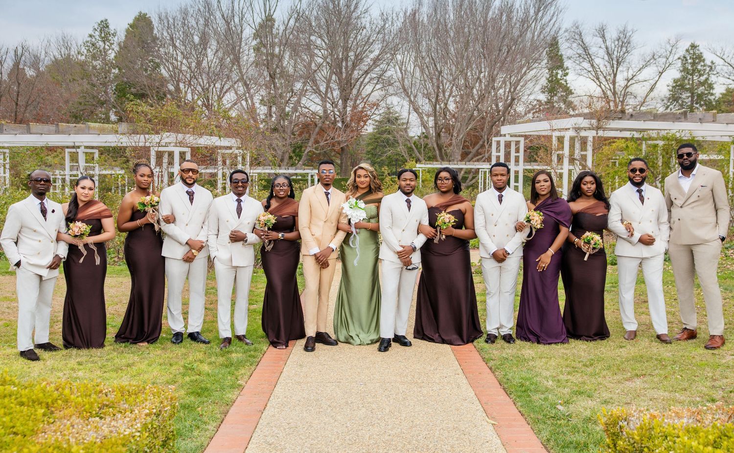 Wedding party poses outdoors in purple bridesmaid dresses and white suits on a brick pathway at a garden venue.