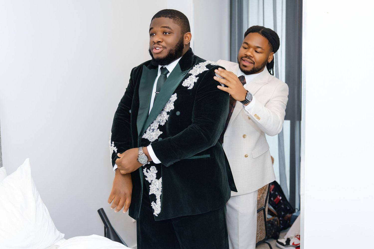 A groom in a green velvet floral suit smiles as a groomsman in white adjusts his jacket.