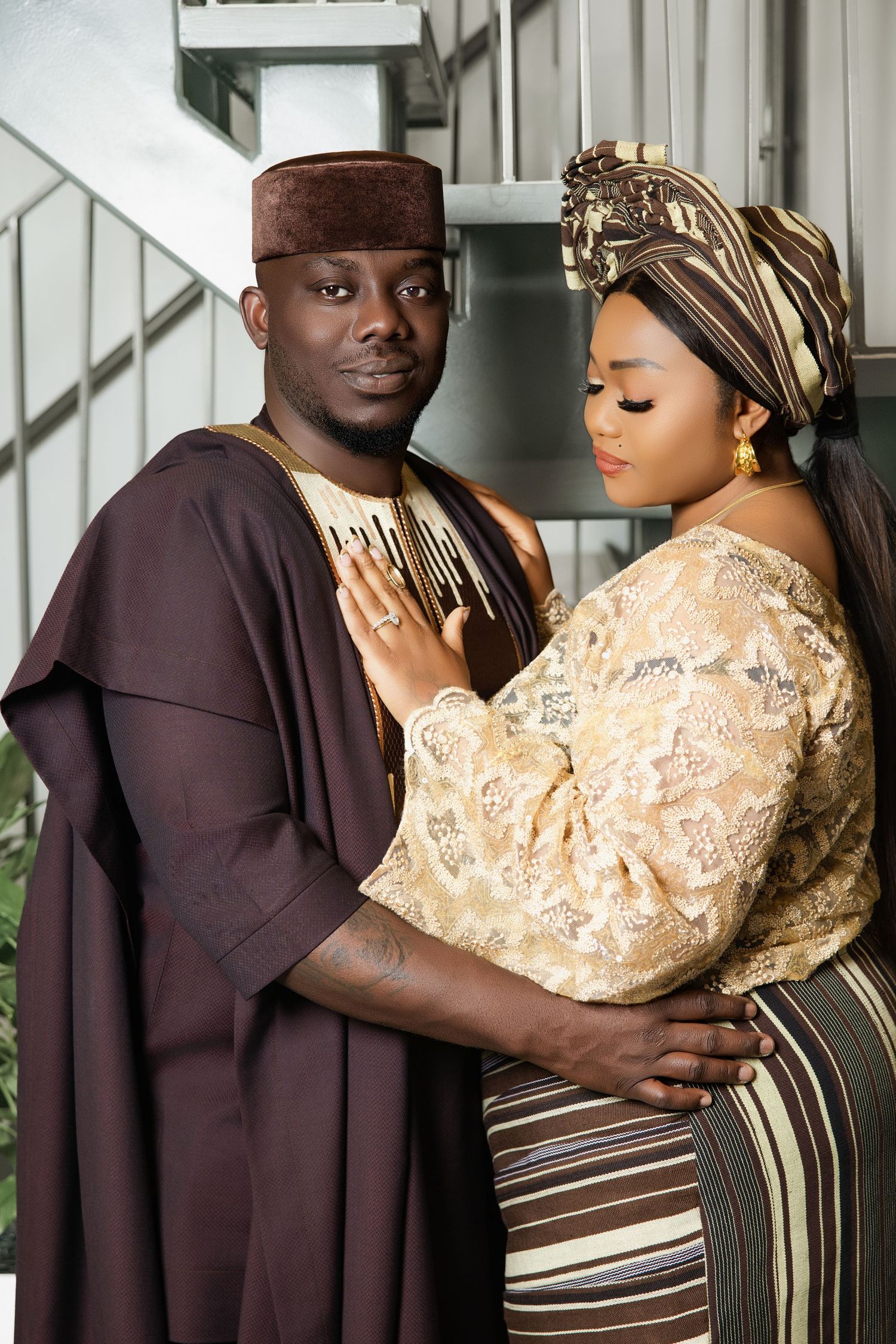 Couple in traditional West African attire, man in brown agbada, woman in gold brocade dress and gele headwrap.