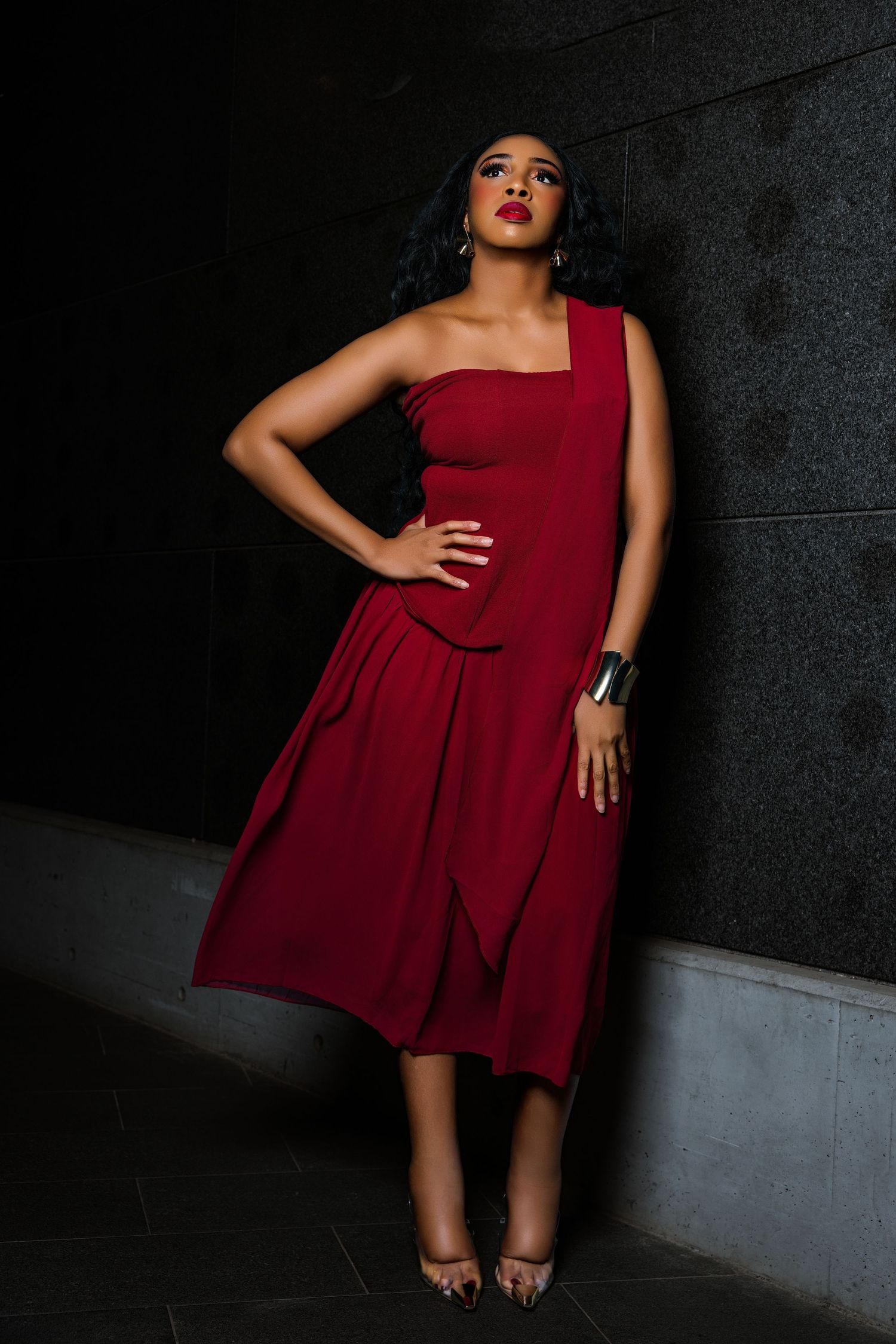 Woman in elegant red one-shoulder midi dress with red lips poses confidently against dark wall.