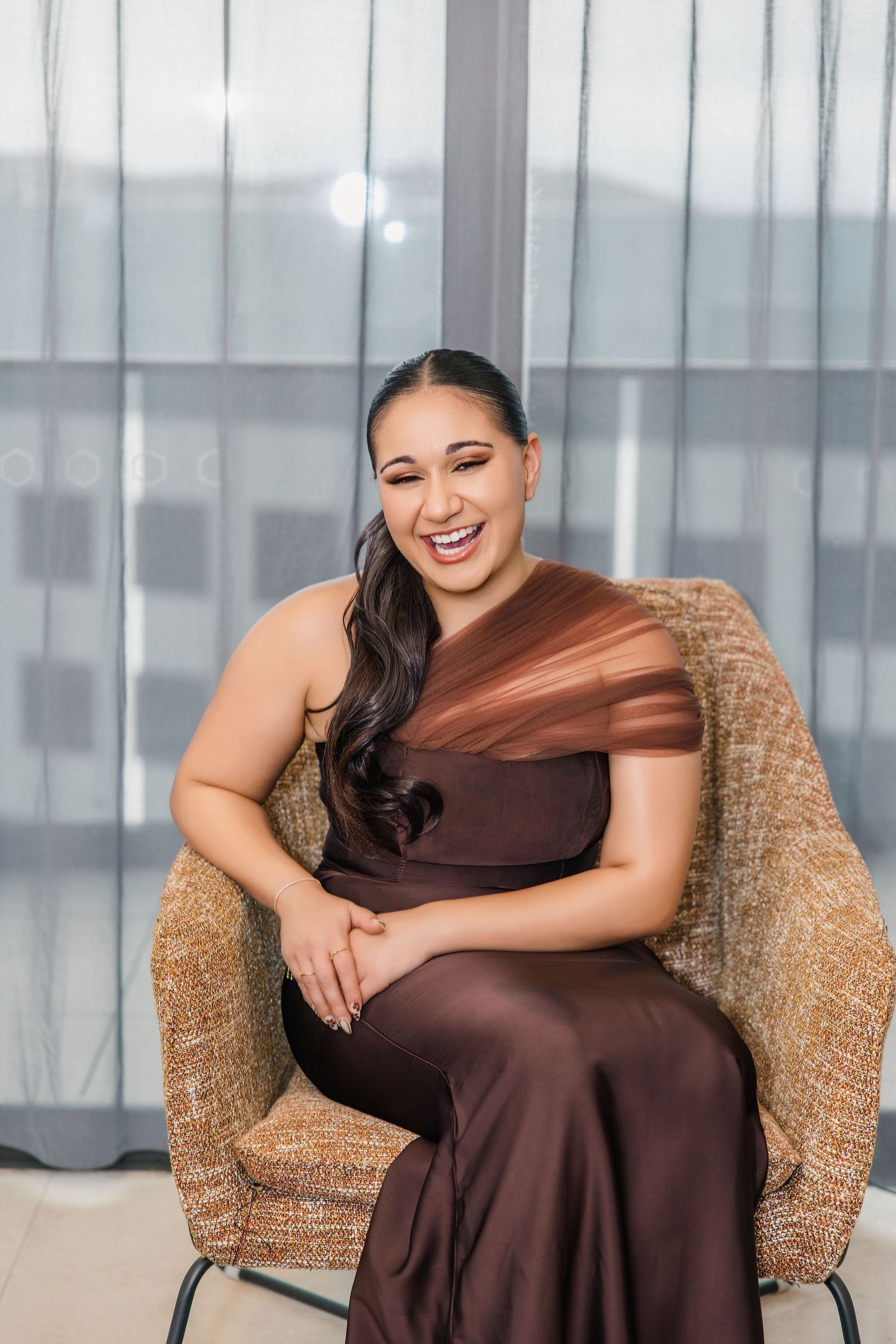 Laughing woman in a brown one-shoulder satin gown seated in a wicker chair by sheer curtains.