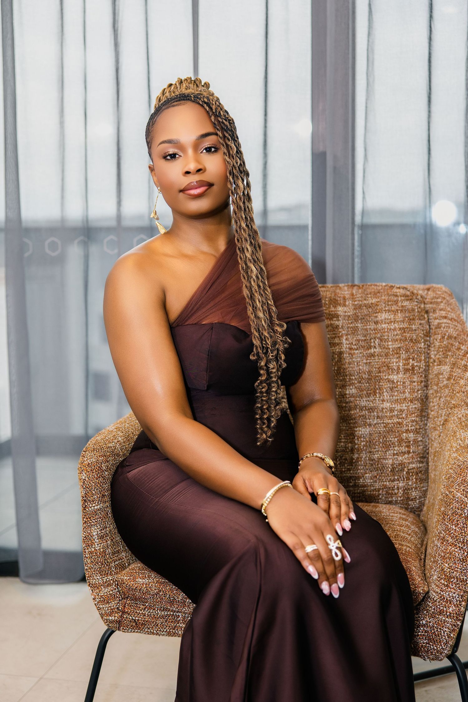A poised woman with braided hair wearing a brown one-shoulder formal gown sits in a wicker chair.