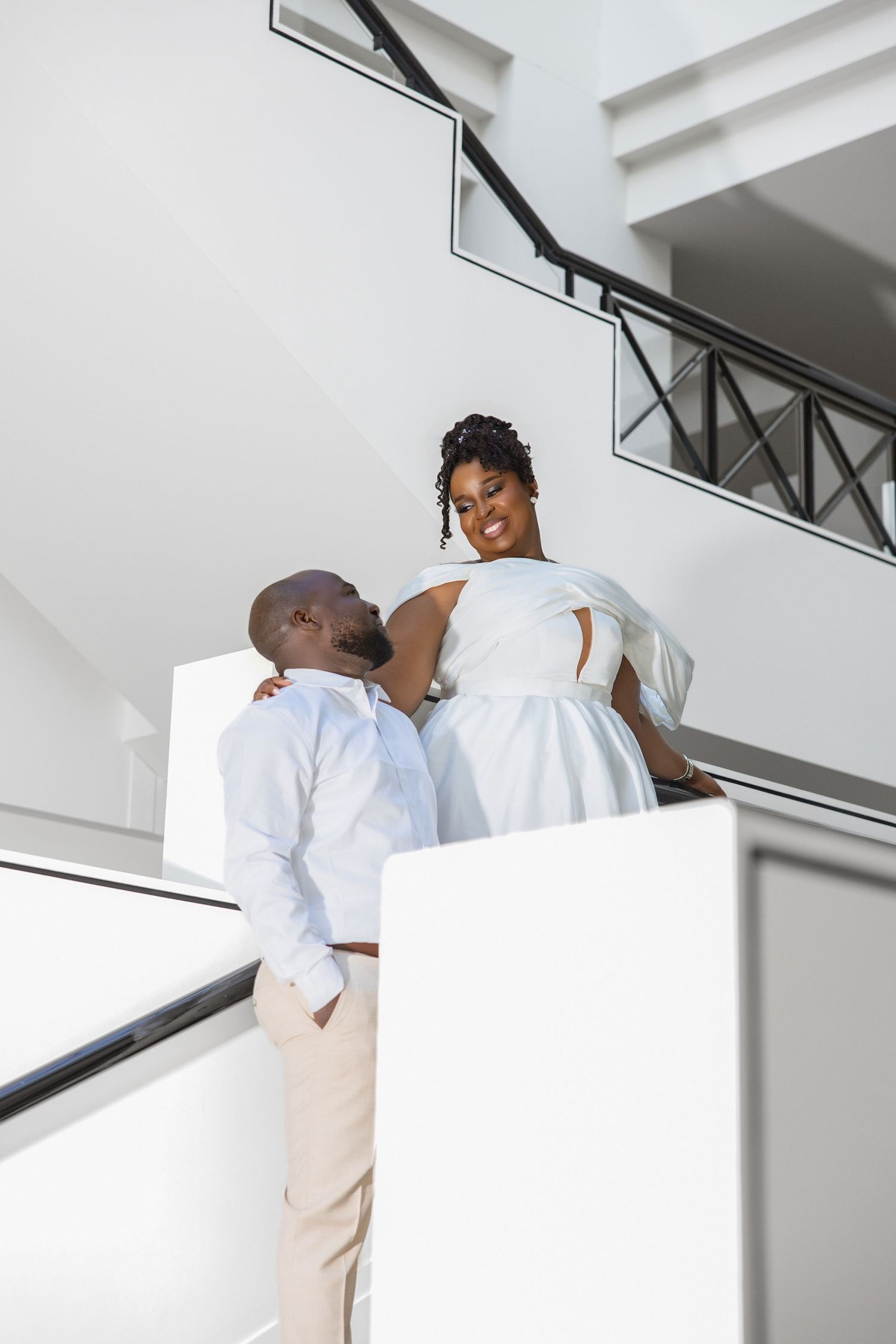 Smiling bride in white dress sitting on staircase railing with groom in light blue shirt looking up at her.