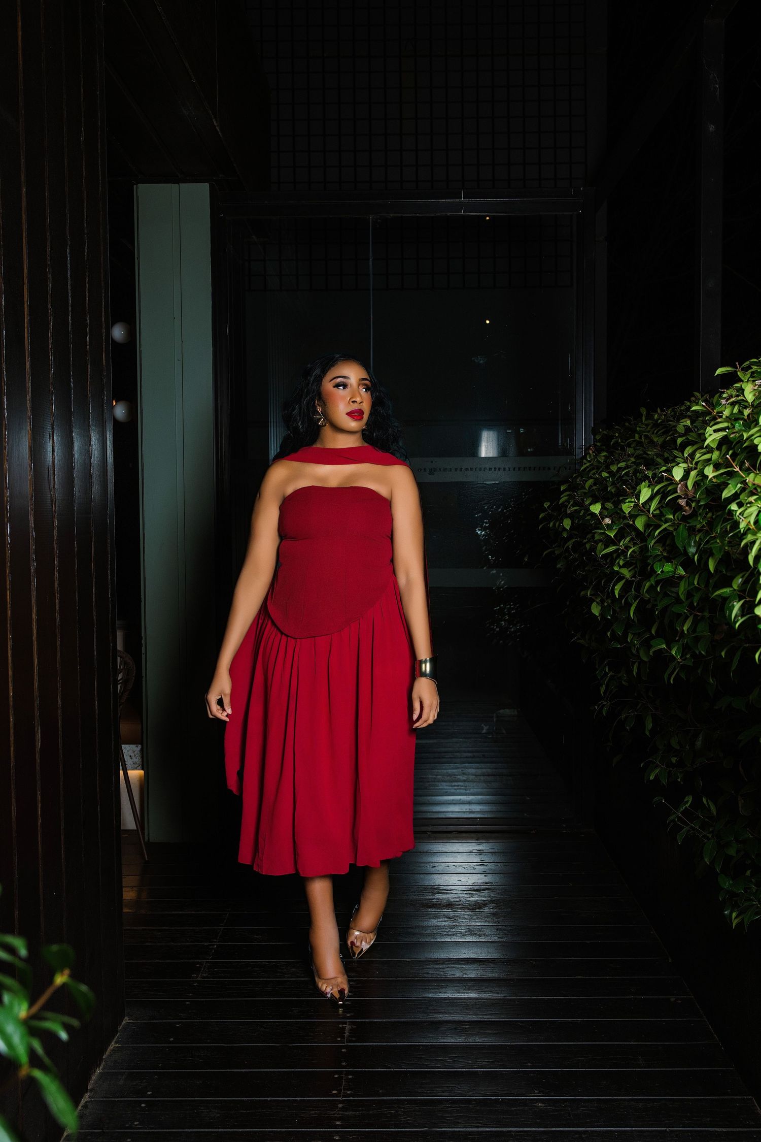 Woman in elegant red strapless midi dress walking down a dark hallway with lush green plants.
