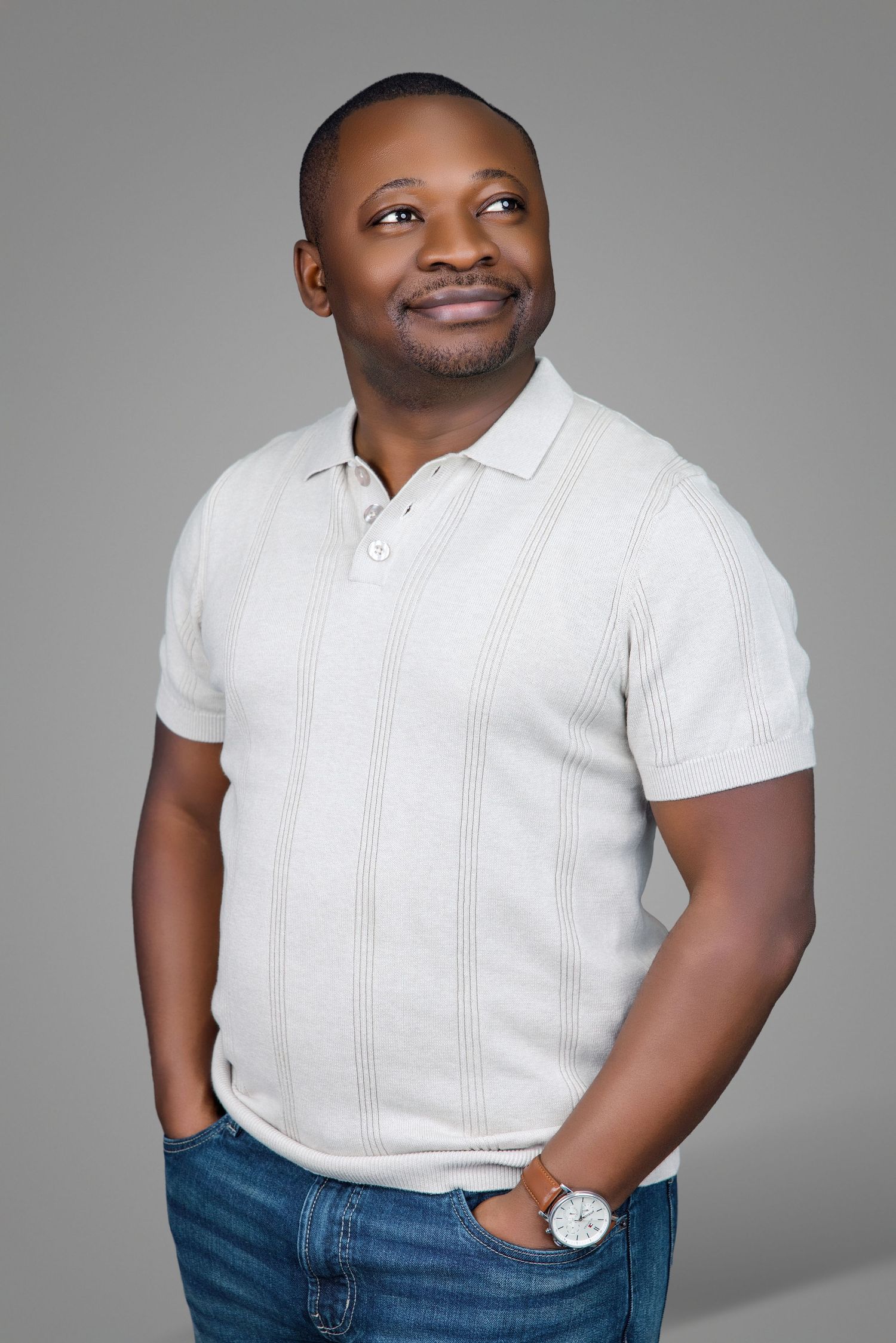 A Black man in a white ribbed polo shirt and jeans smiles upward while posing against a neutral gray background.