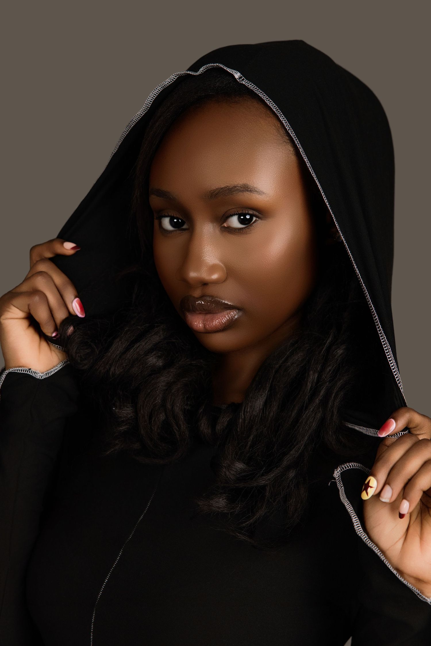 Young Black woman in black hooded zip-up dress holding hood up, studio portrait on gray background.