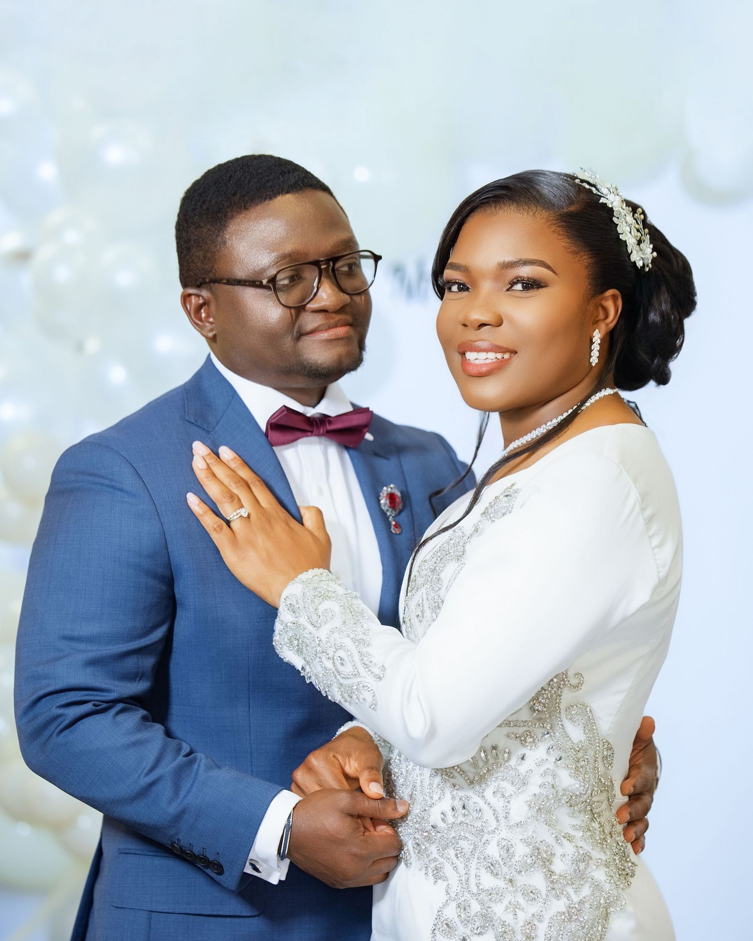 Elegant couple in wedding attire, groom in blue suit with bow tie, bride in white lace gown with hair accessory.