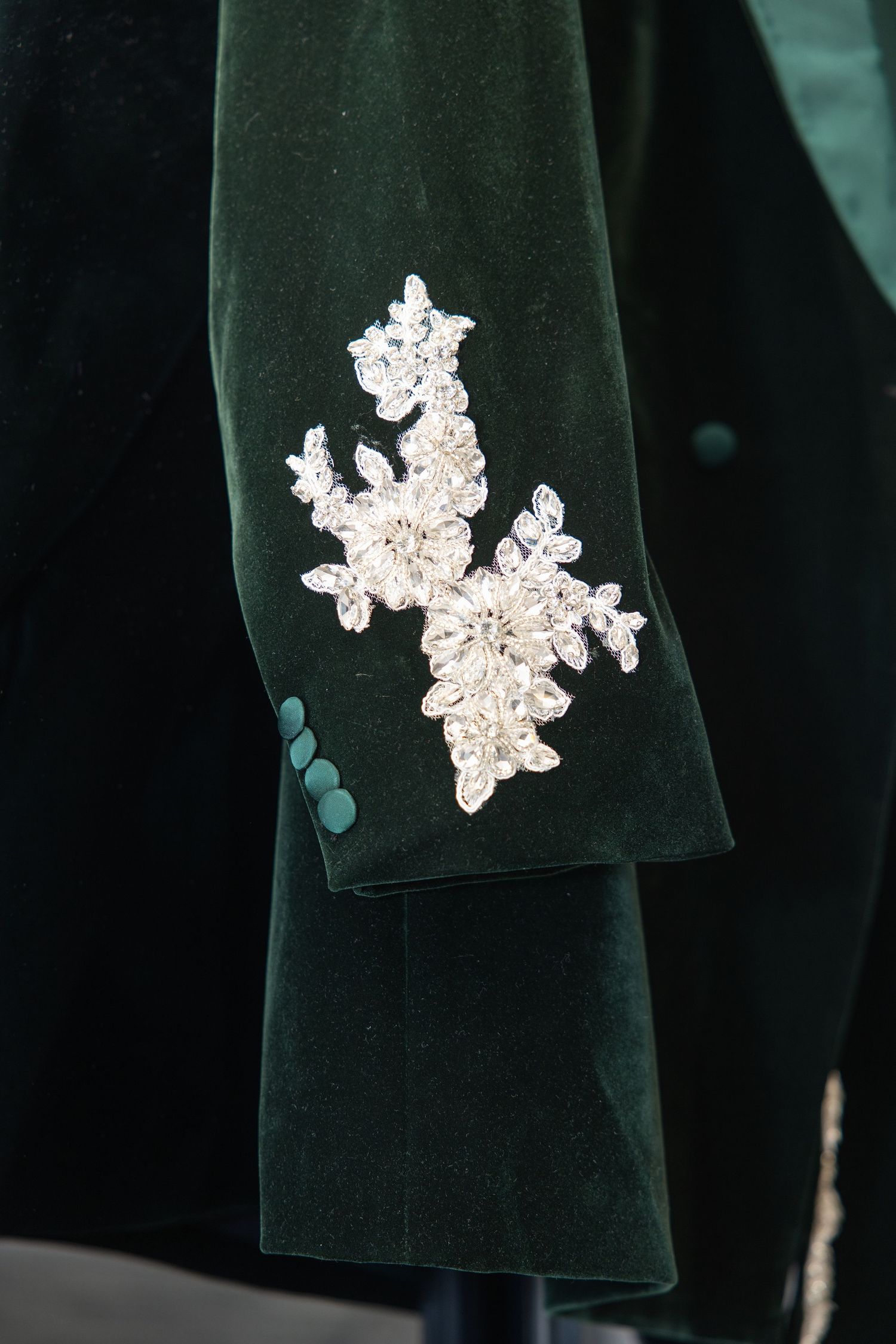 Close-up detail of white crystal floral applique embroidery on a dark green velvet formal jacket sleeve.