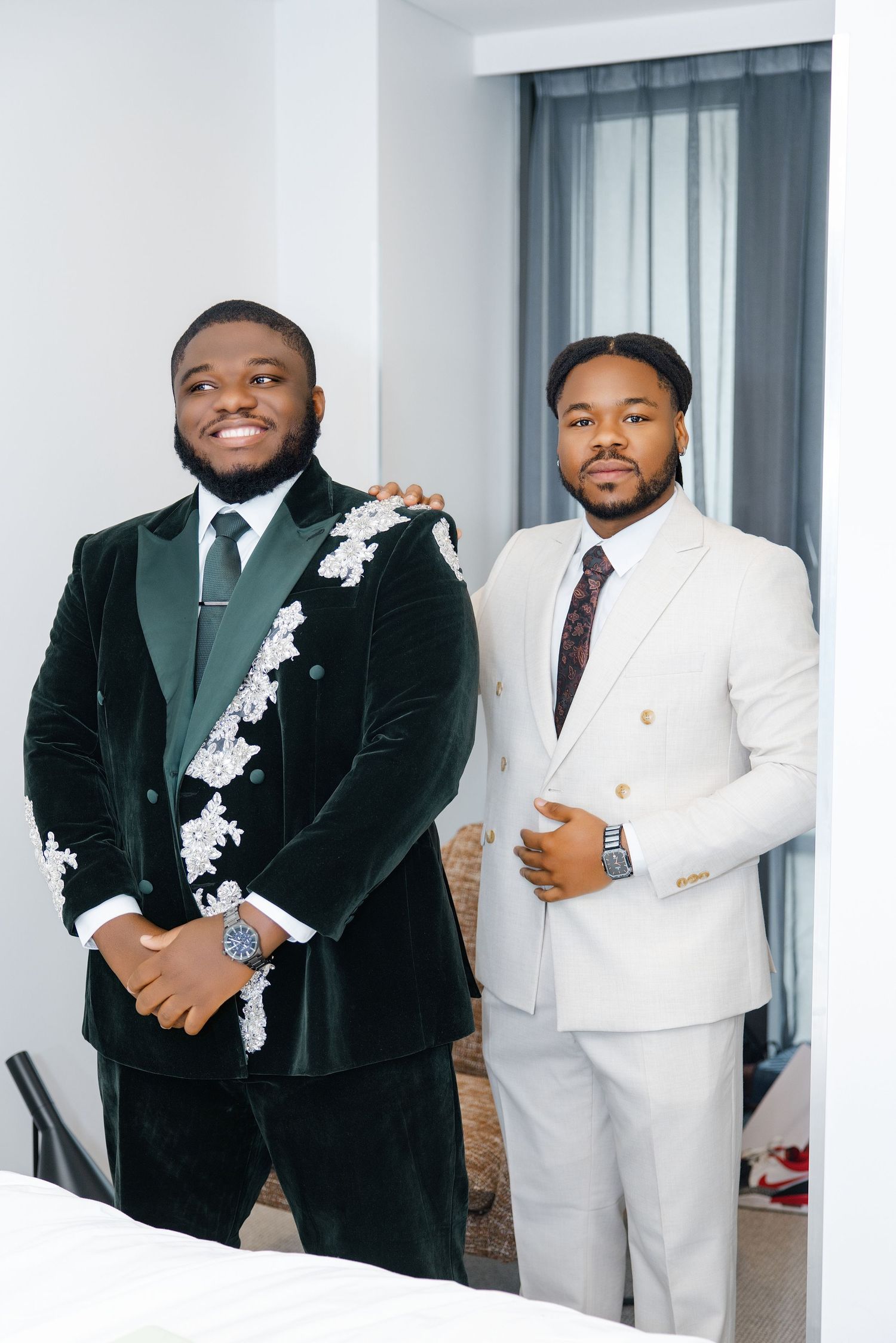 Two well-dressed men posing in elegant suits, one in green velvet with floral embroidery, the other in a cream double-breasted suit.