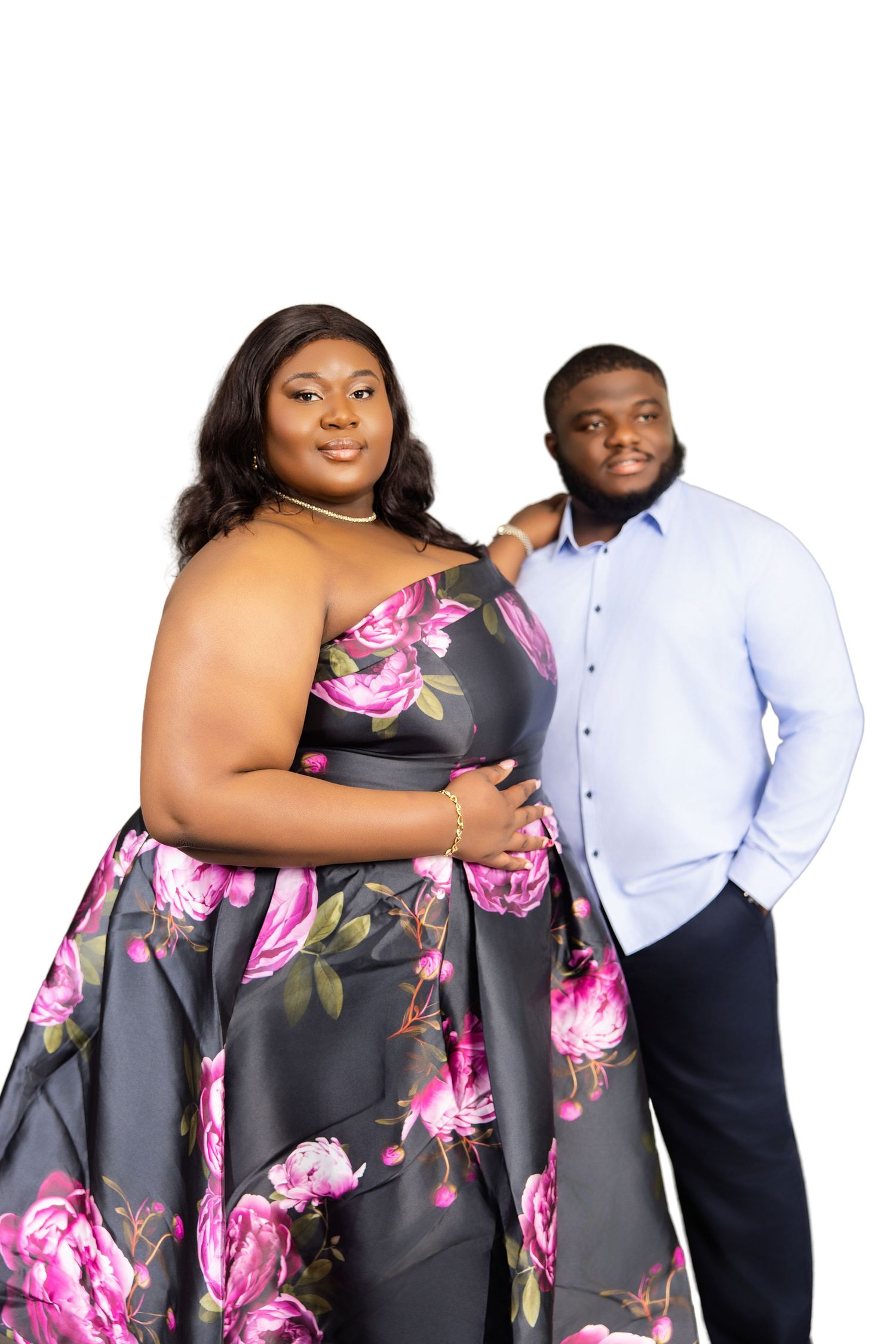 A couple poses elegantly, woman in a floral gown and man in a blue shirt, against white background.