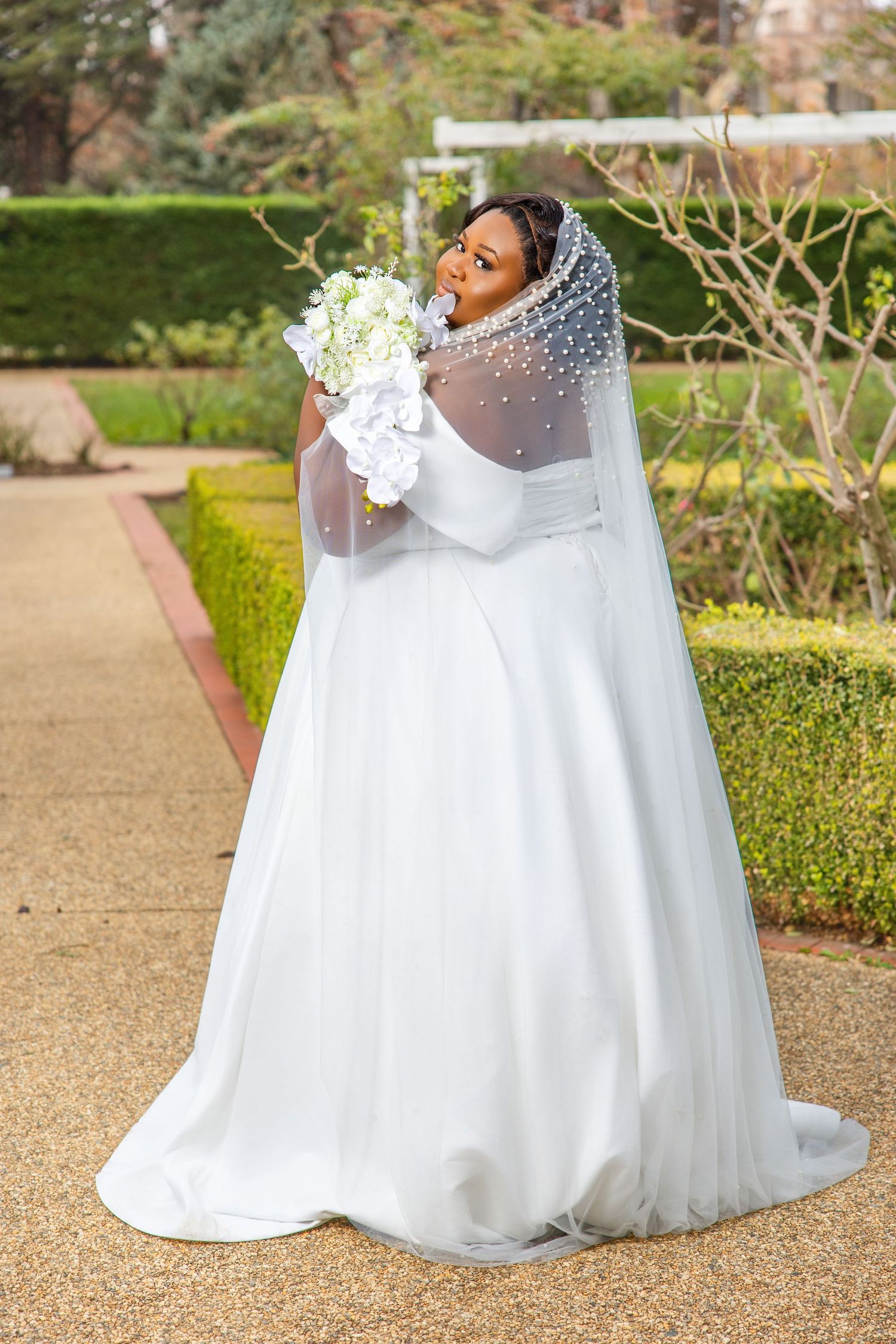 Elegant plus-size bride in white ball gown with pearl-embellished veil holding white bouquet in garden.