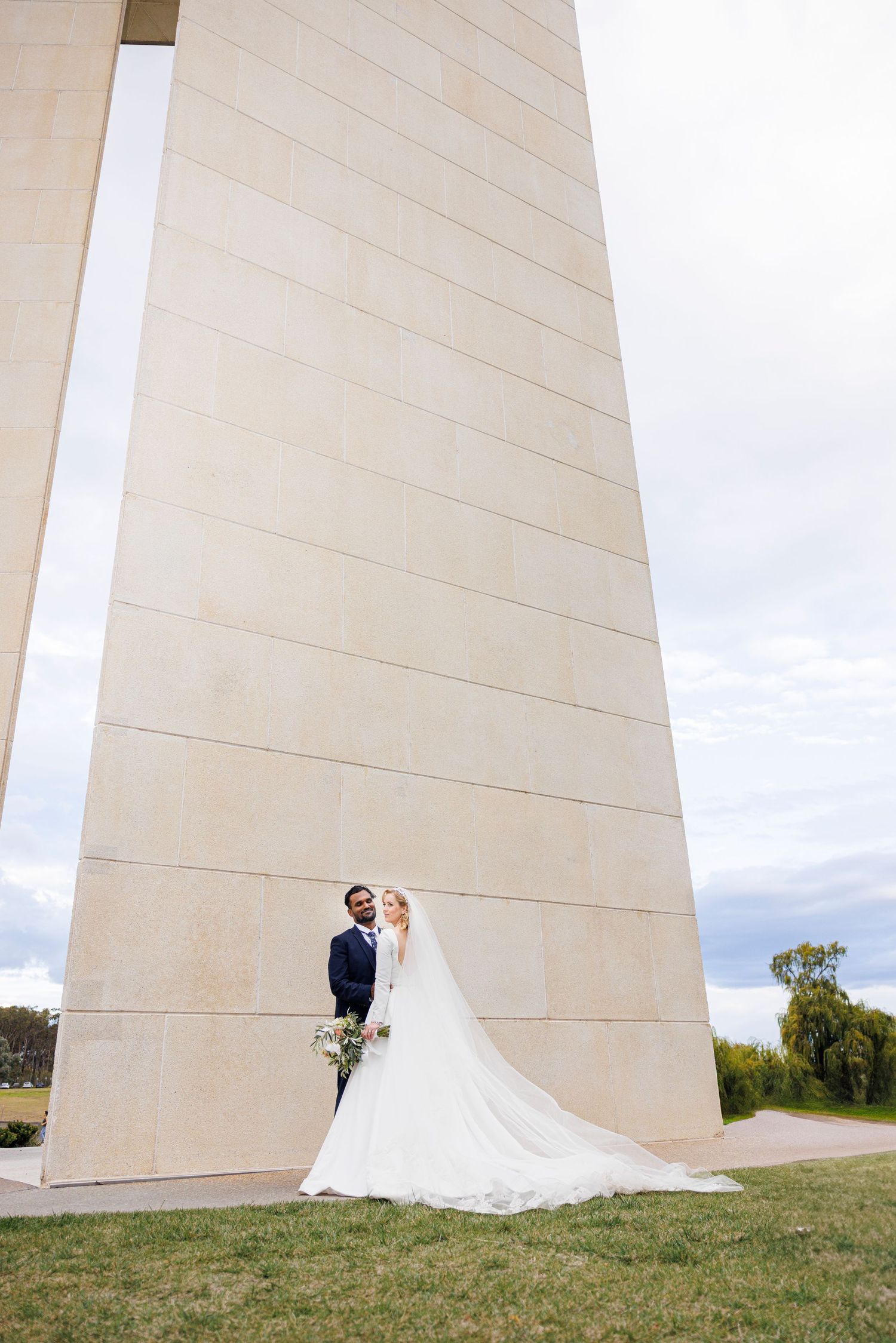 Bride and groom standing together beneath a tall stone monument on their wedding day.