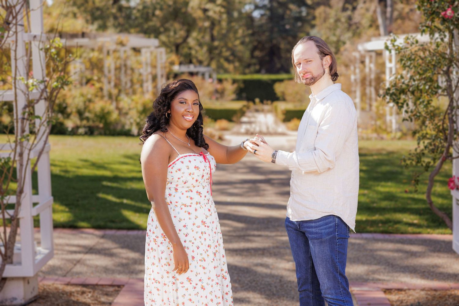 A smiling couple stands together near a white arbor in a lush garden, sharing a cheerful moment.
