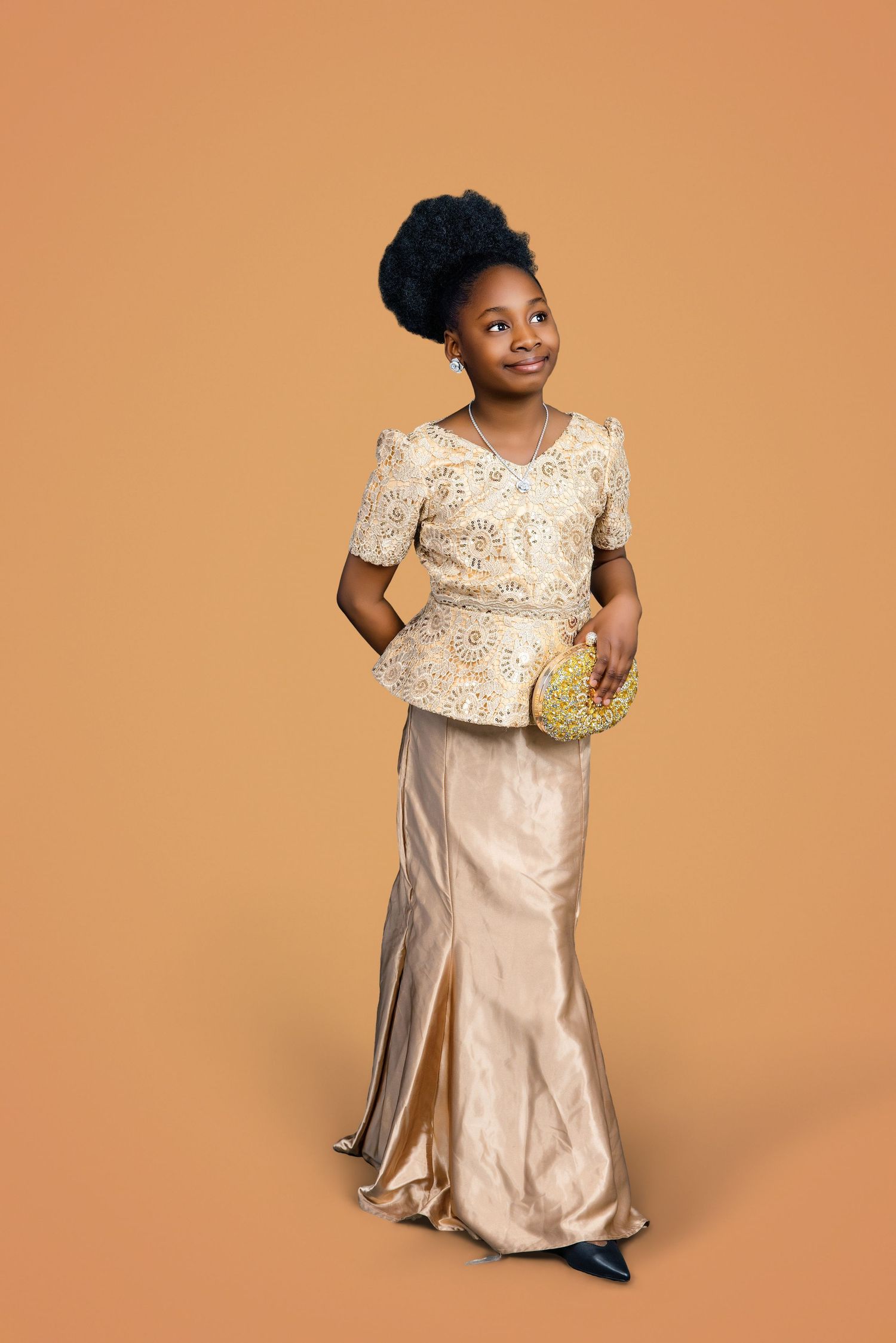 Young Black girl in elegant gold lace outfit holding a decorative clutch against a warm beige background.