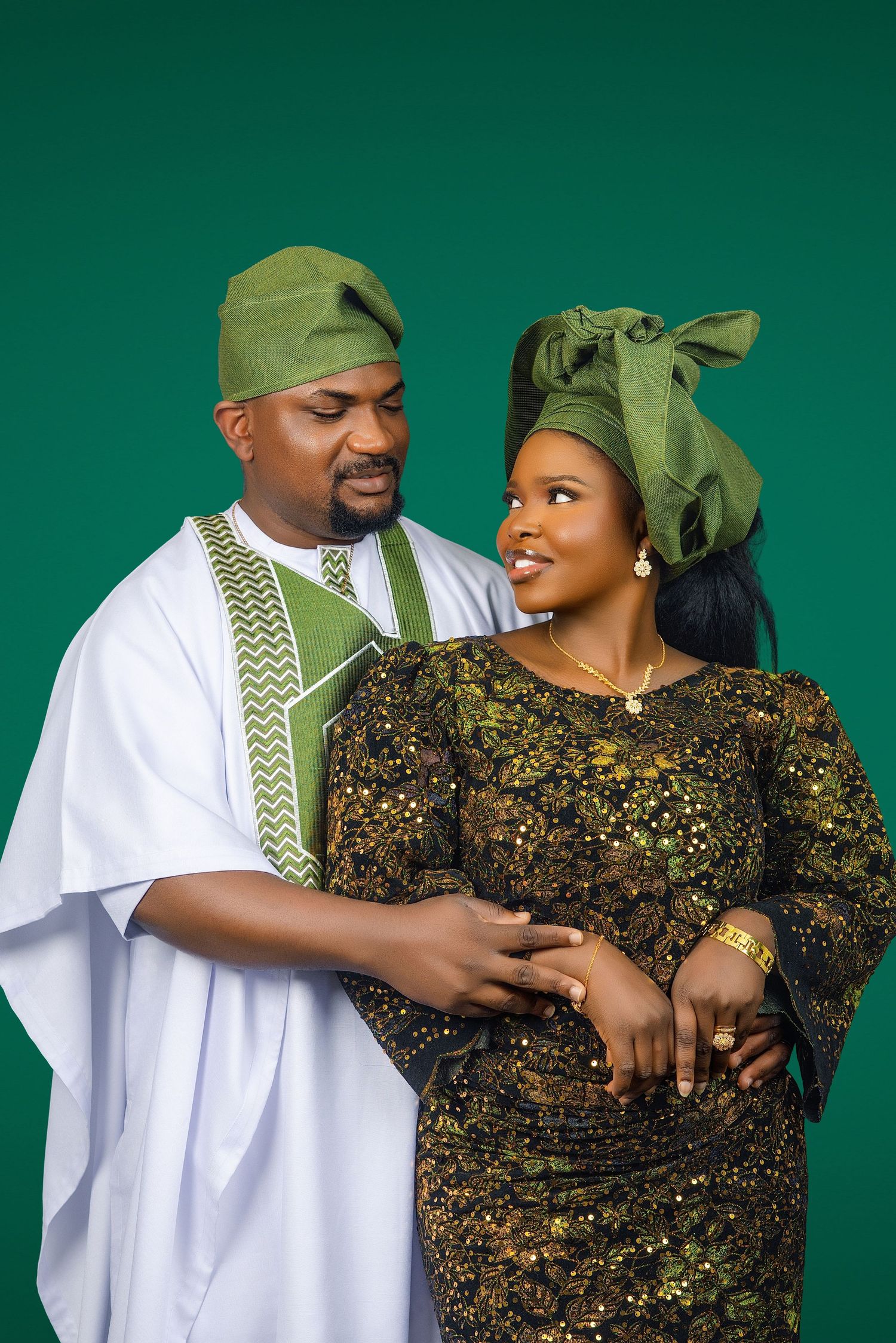 Nigerian couple in traditional green and white Yoruba attire posing together against a teal studio background.