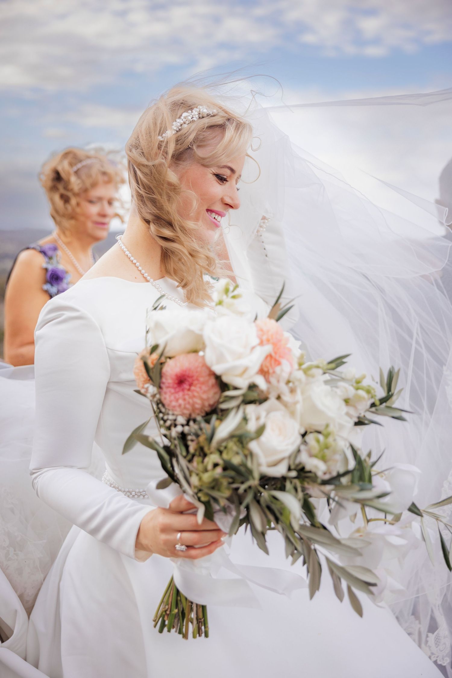 Smiling bride in white gown holding pink and white bouquet with veil flowing in the wind.