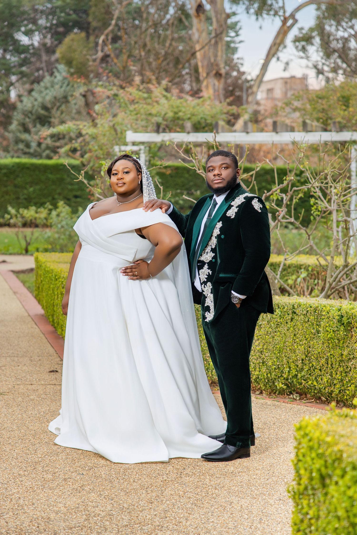 Bride in white off-shoulder gown and groom in green velvet floral blazer posing together in garden.