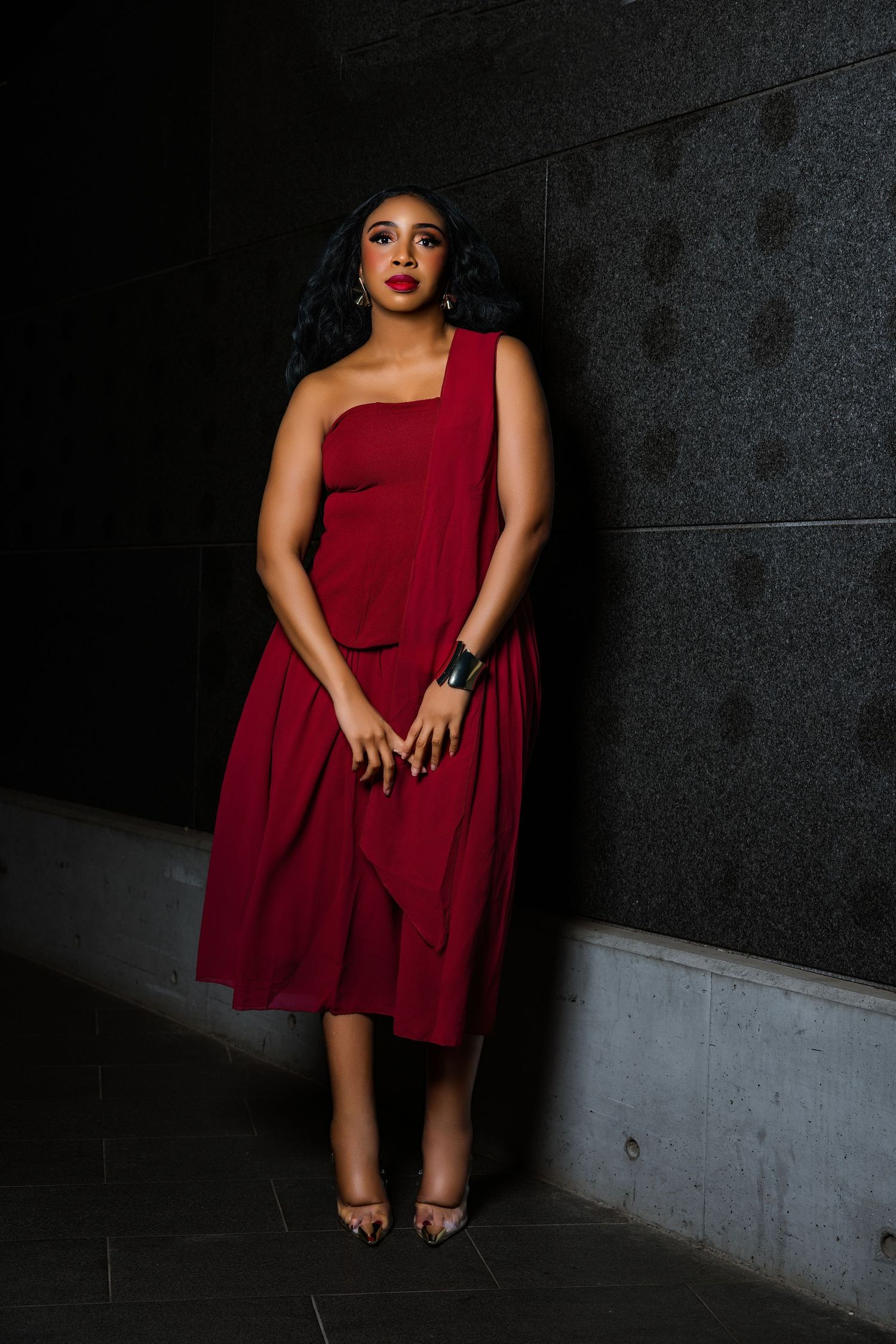 A woman in a red one-shoulder midi dress poses elegantly against a dark granite wall wearing strappy heels.