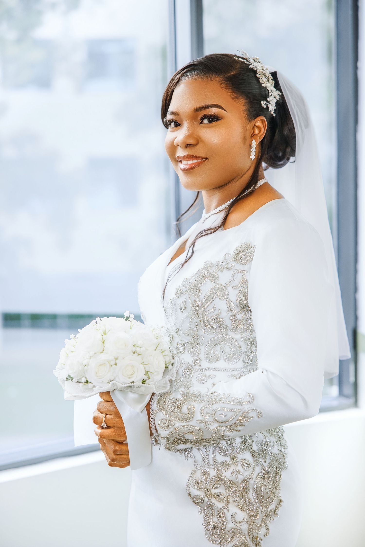 Smiling Black bride in white long-sleeve beaded gown holding white rose bouquet, wearing veil and pearl accessories.