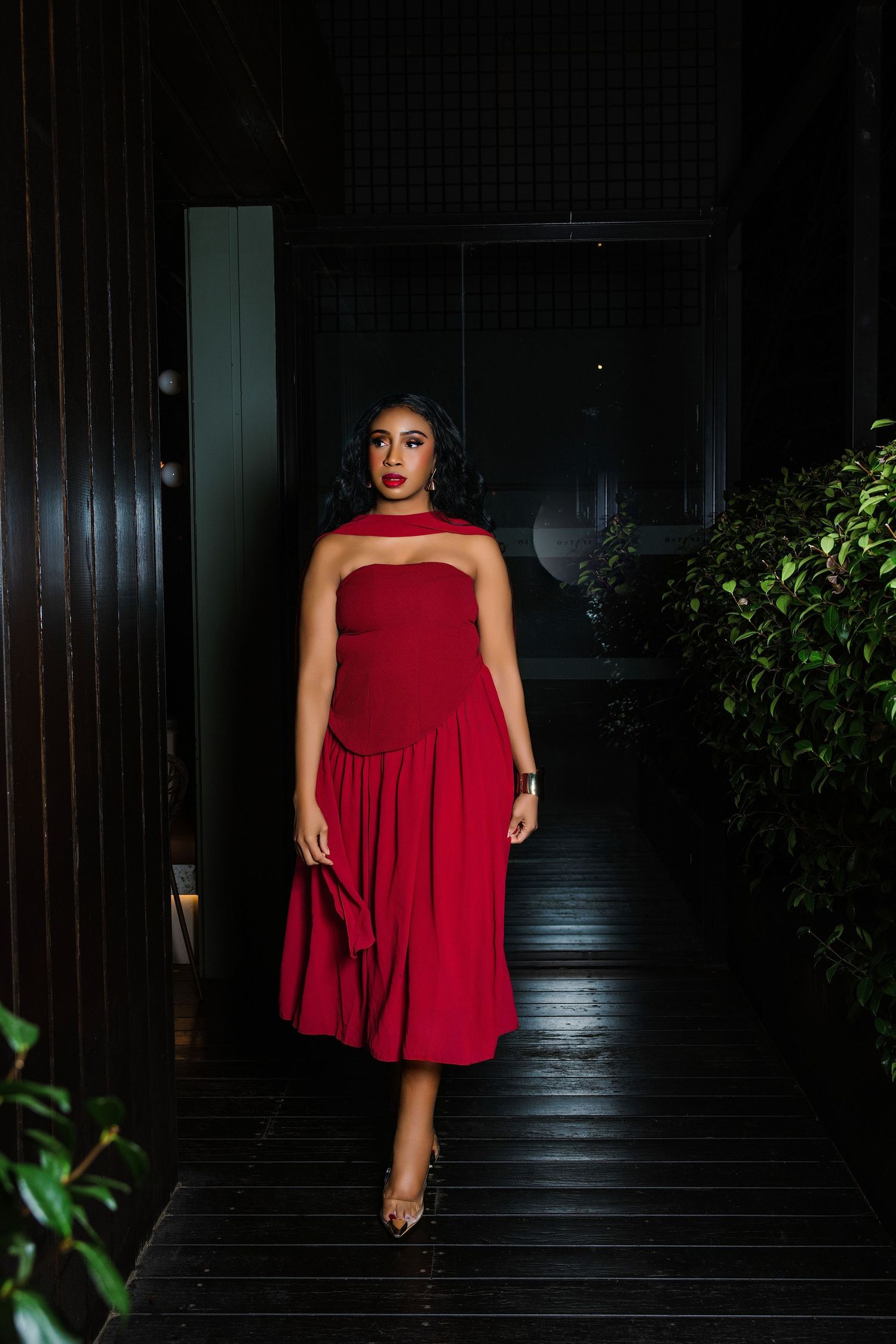 Woman in elegant red midi dress with halter neckline poses in dimly lit corridor with dark wood and greenery.