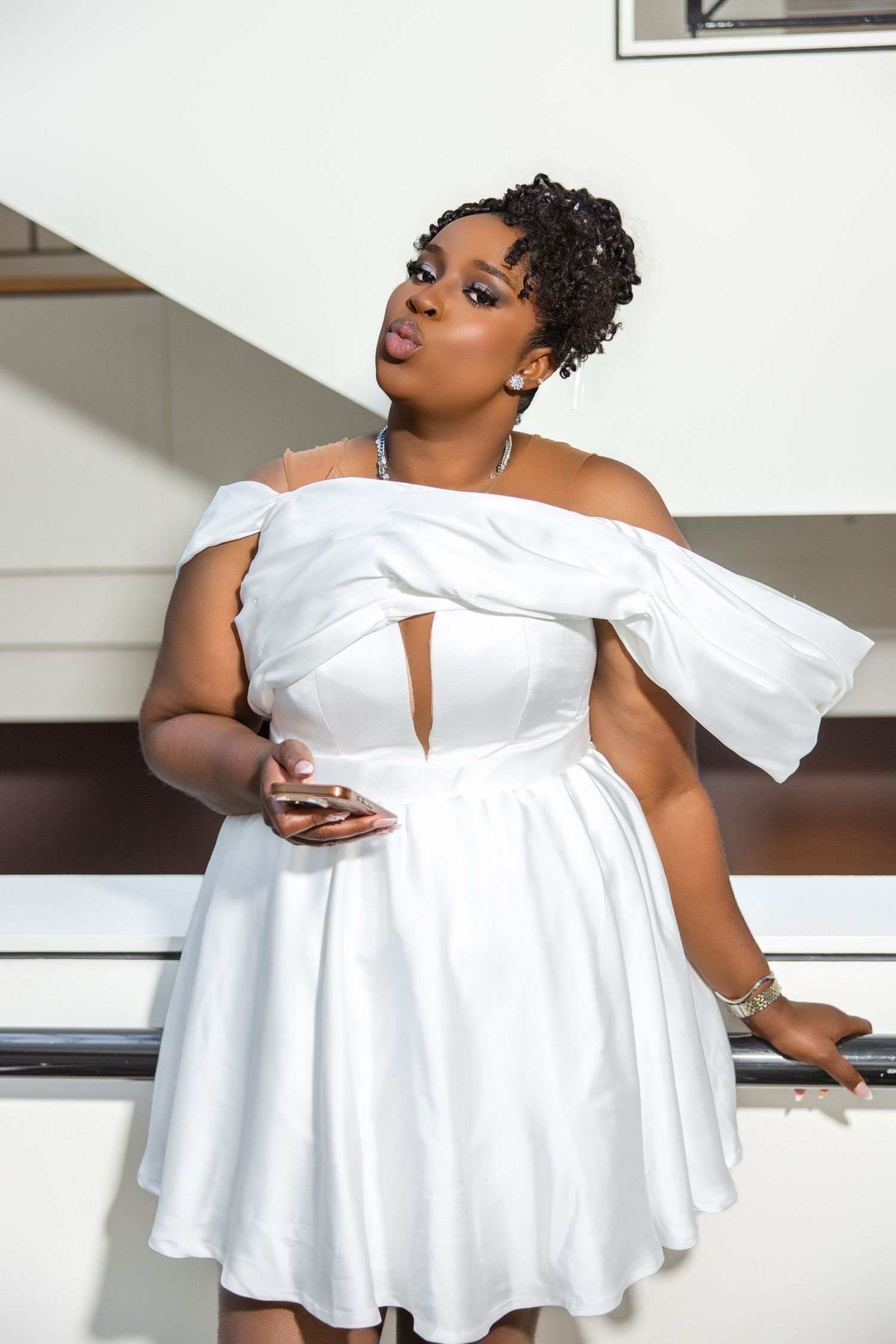 Bride in white off-shoulder mini dress with cutout detail posing confidently on modern white staircase.