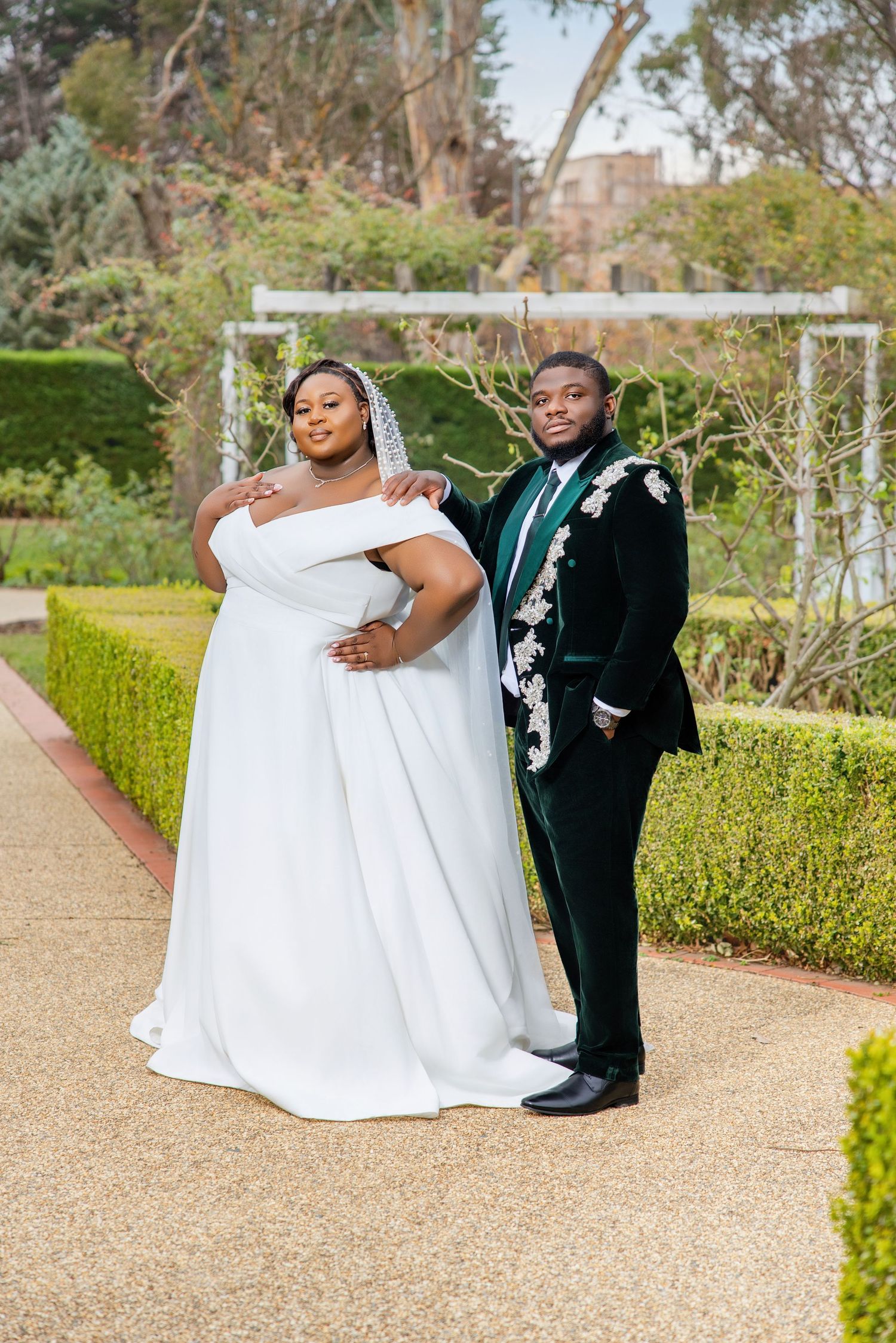 A bride in a white gown and groom in a dark green floral tuxedo pose elegantly in a garden setting.