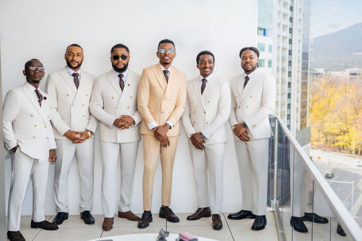 Six well-dressed groomsmen in beige and white suits pose together on a balcony with a city skyline backdrop.