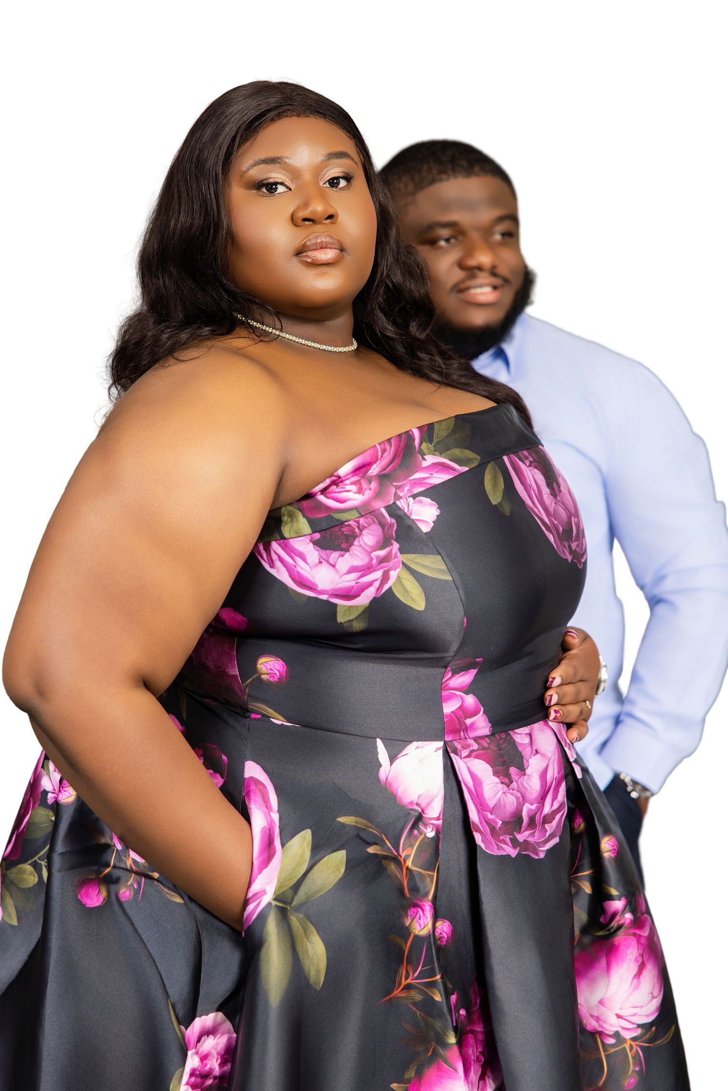 A plus-size couple poses formally, woman in black floral gown and man in blue dress shirt against white background.