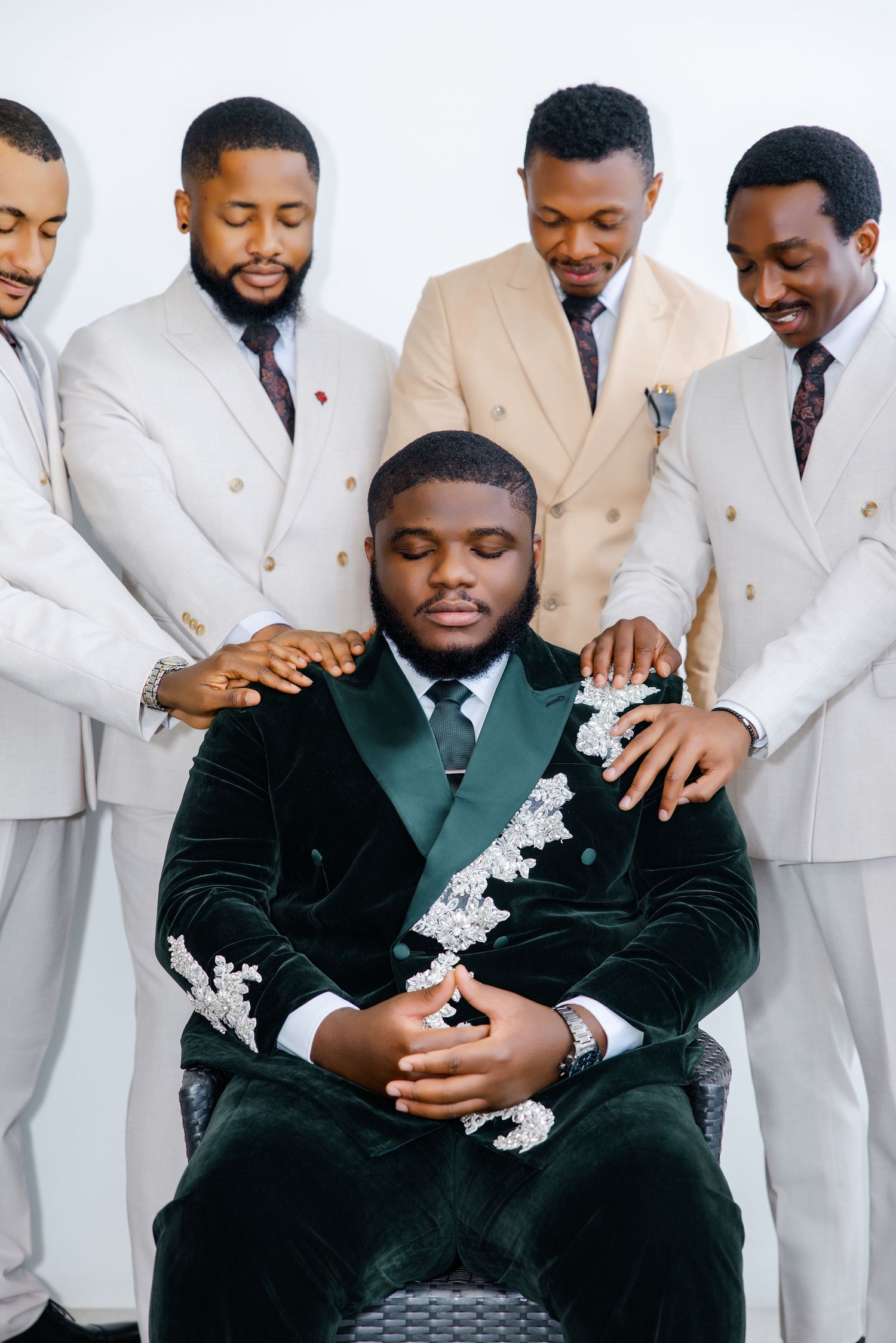 Four groomsmen in beige suits lay hands on a seated groom in a green velvet embroidered suit.