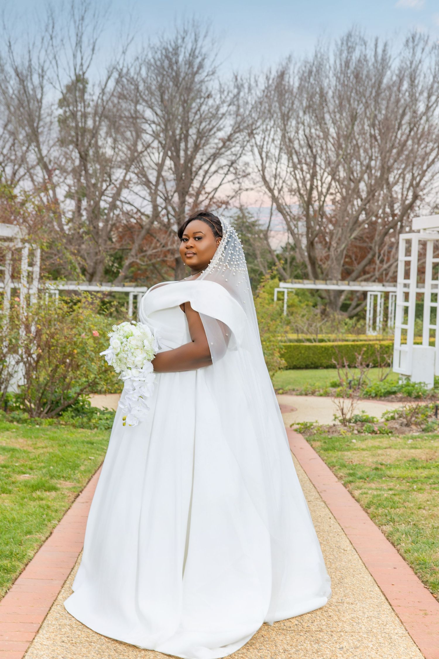 A bride in a white off-shoulder gown with a long veil holds a bouquet in a garden pathway.