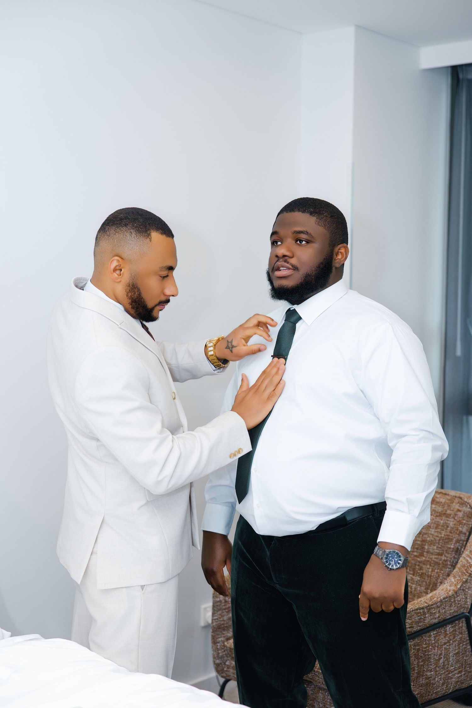 A man in a white suit helps a groom in a white shirt adjust his green tie before the wedding ceremony.