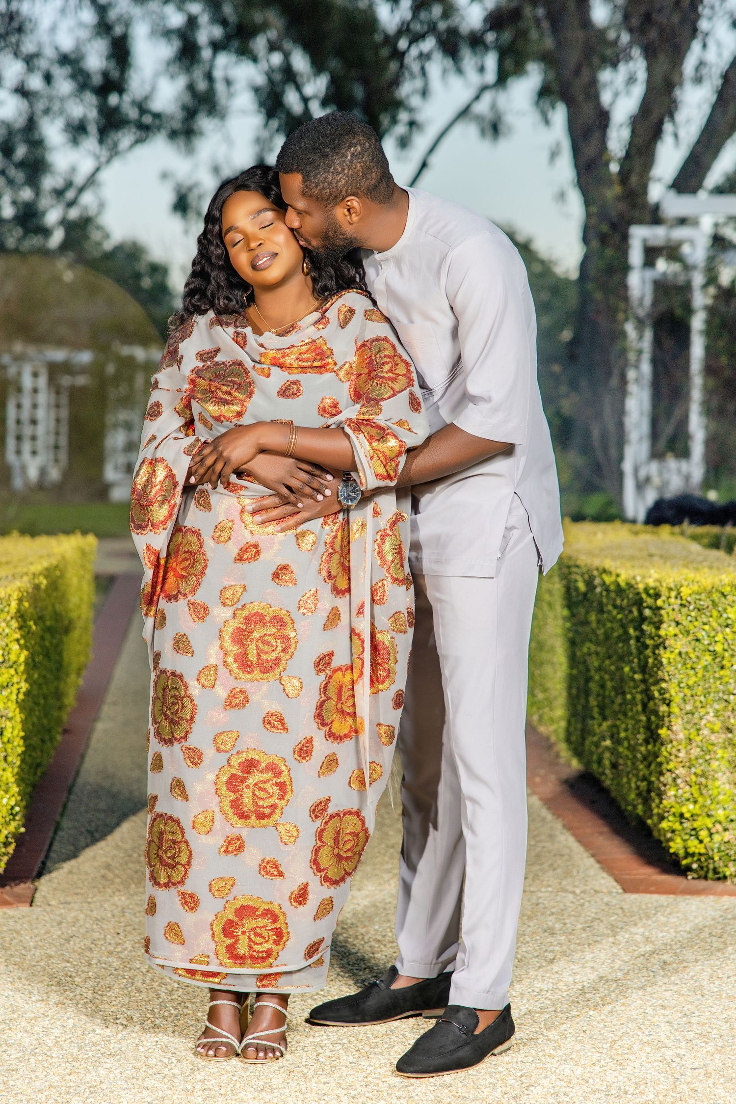 A man kisses a pregnant woman wearing a floral dress as they pose together in a lush garden setting.