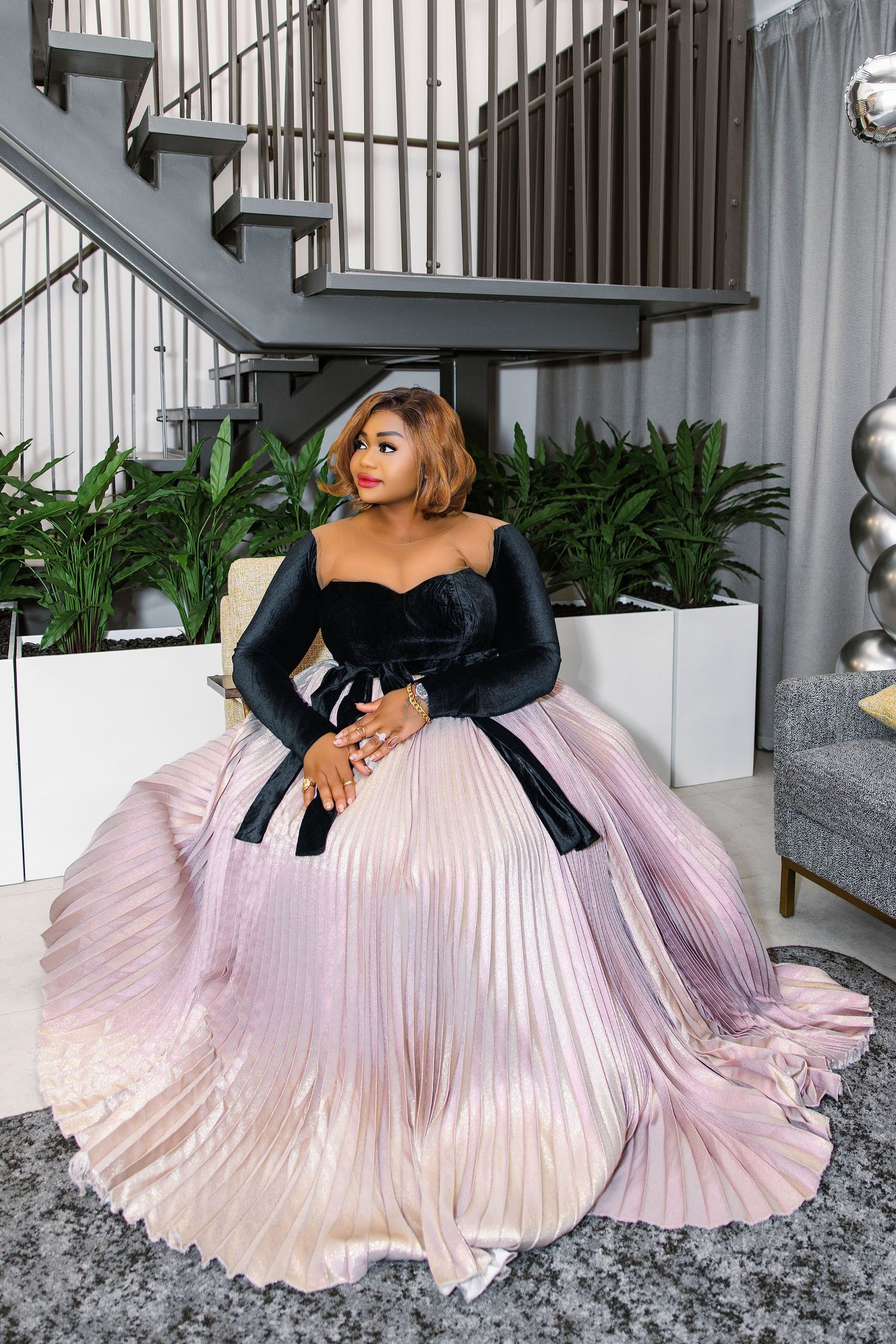 Woman in elegant black bodice and pleated lavender ball gown poses gracefully near staircase with lush greenery.