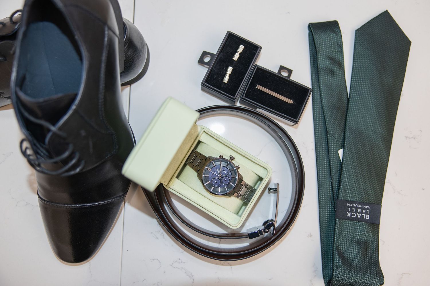 Flat lay of men's formal accessories including black dress shoes, green tie, watch, belt, and cufflinks.
