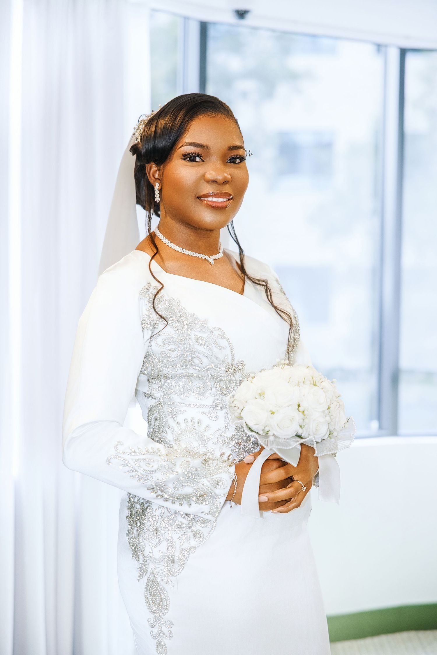 Beautiful Black bride smiling in white long-sleeve lace bridal gown, holding white flower bouquet near bright window.