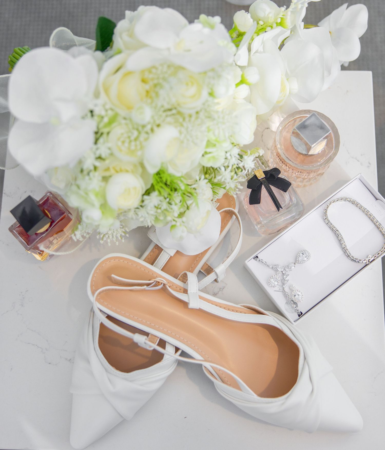 Elegant bridal flat lay featuring white heels, floral bouquet, perfume bottles, and diamond jewelry.