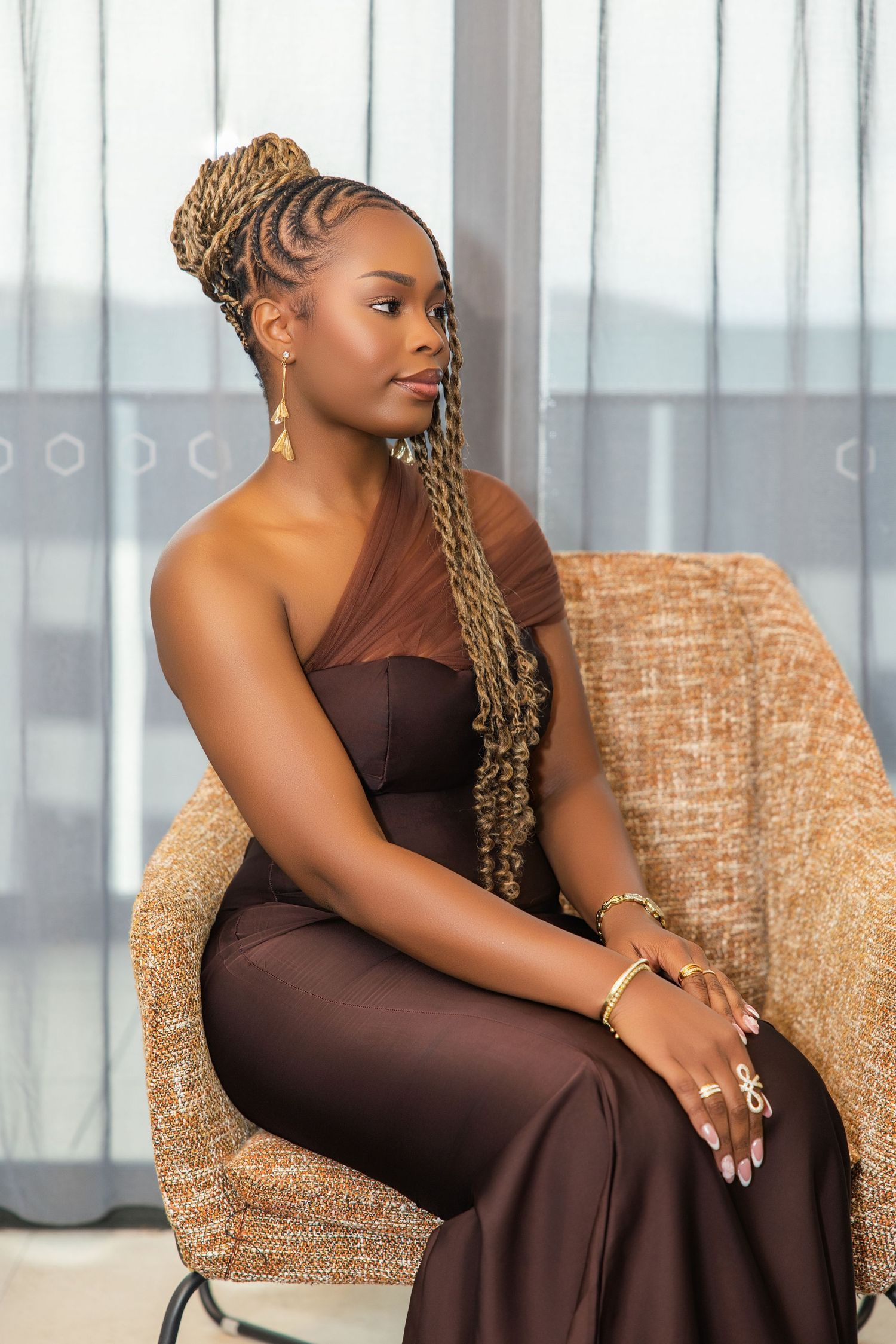 A woman with long braided hair in a brown one-shoulder formal jumpsuit sits gracefully in a wicker chair.