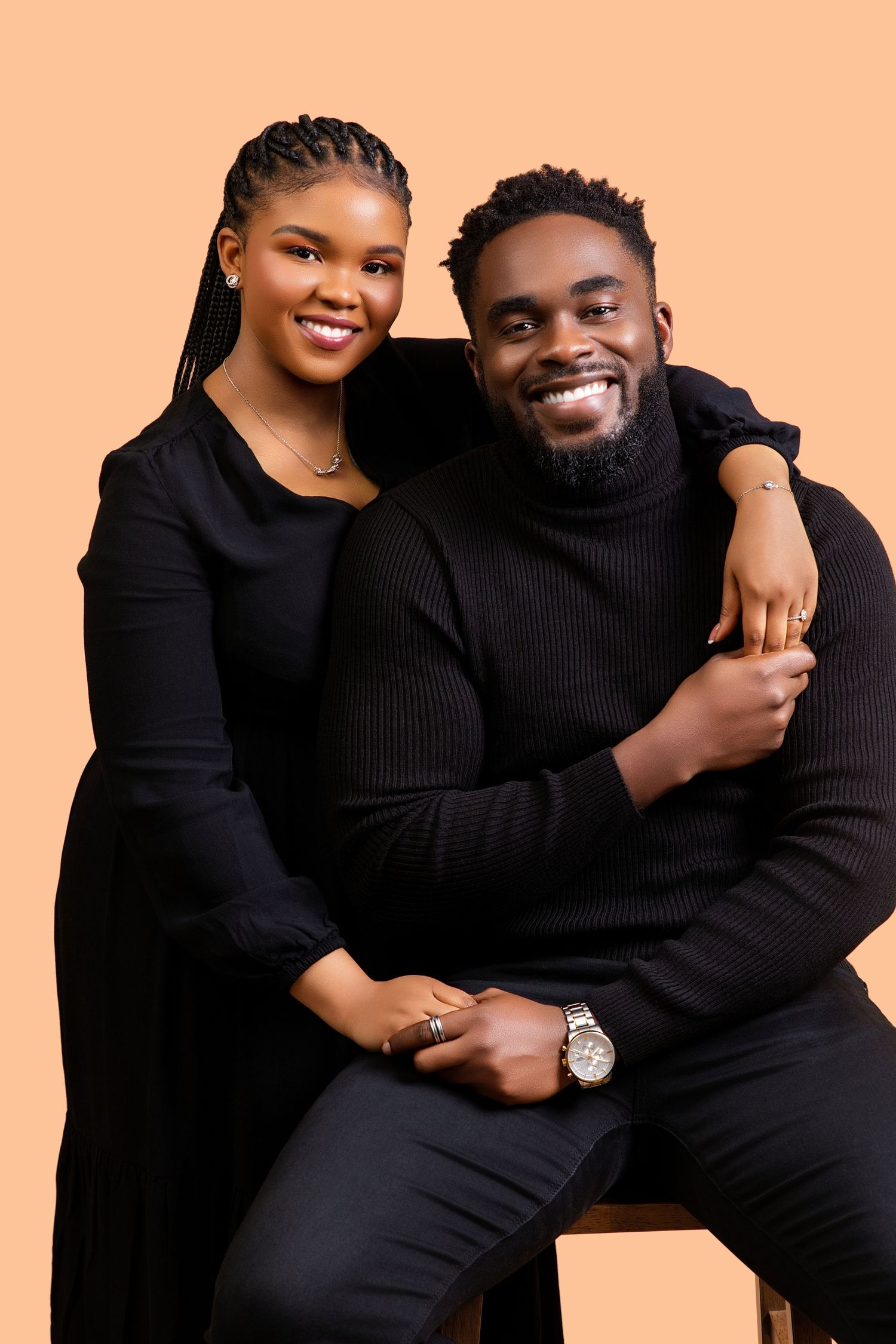 Smiling Black couple in matching black outfits posing together against a warm beige background.